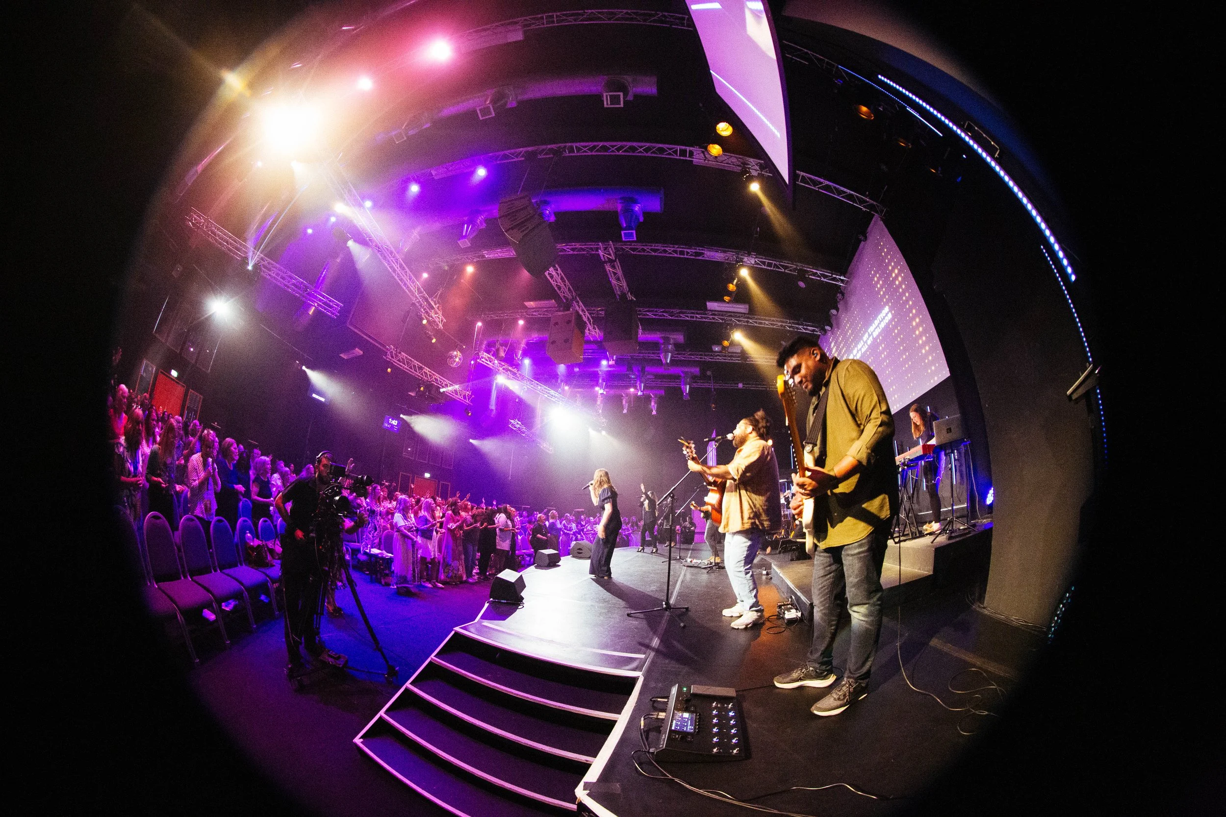 A band performing live on stage at a concert with purple and yellow stage lighting, and an audience watching.