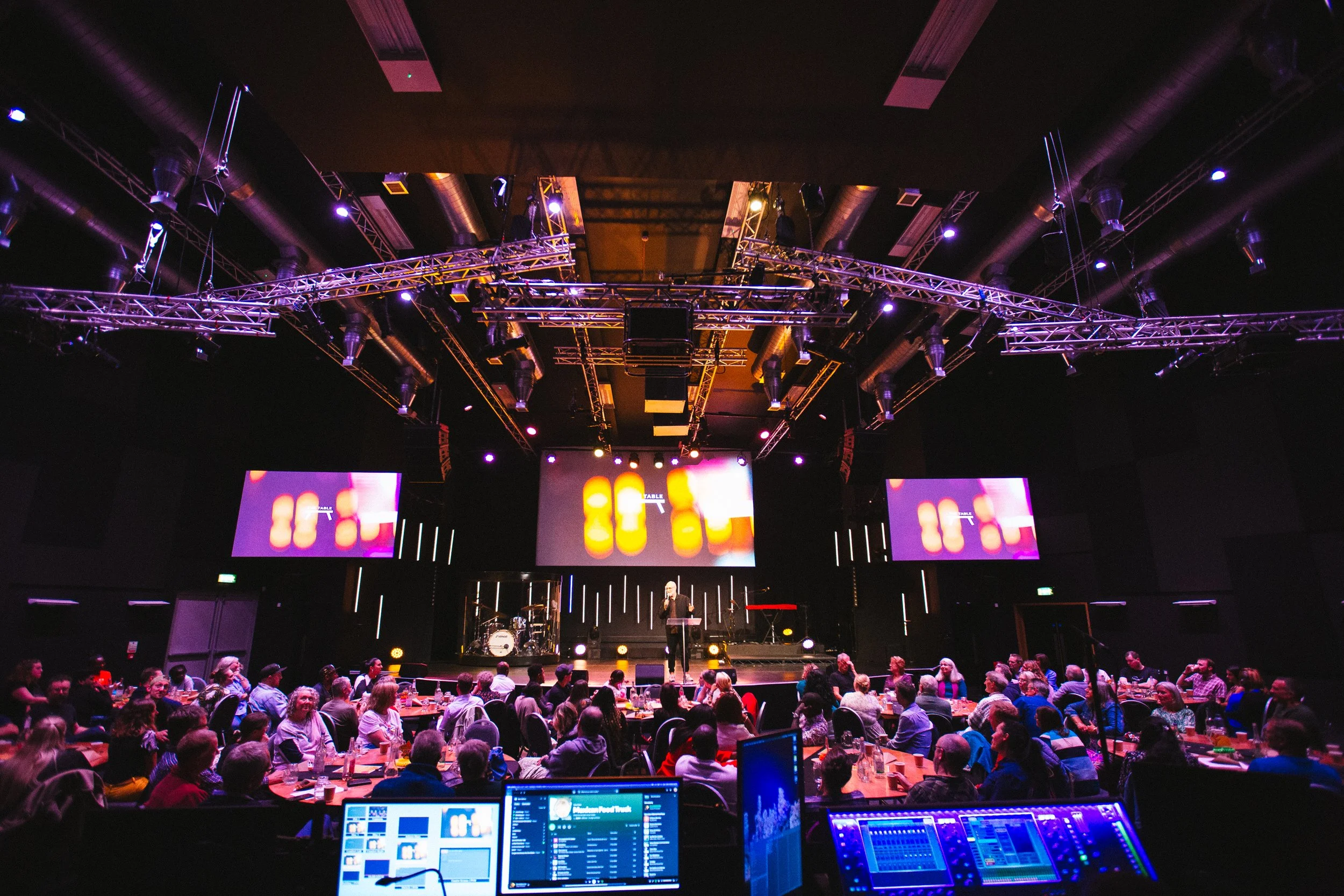An indoor concert or event venue with a stage, microphones, drum set, and large screens displaying colorful graphics. Audience seated at tables facing the stage, with sound and light equipment visible in the foreground.