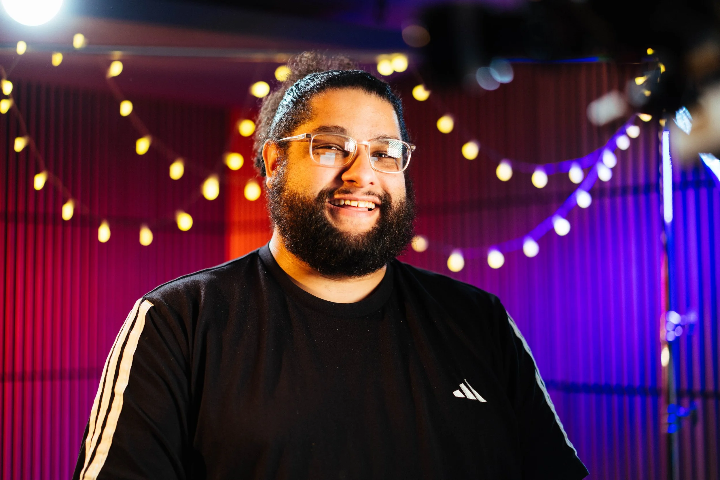 Man with glasses and a beard smiling in front of colorful string lights and a red and purple backdrop.