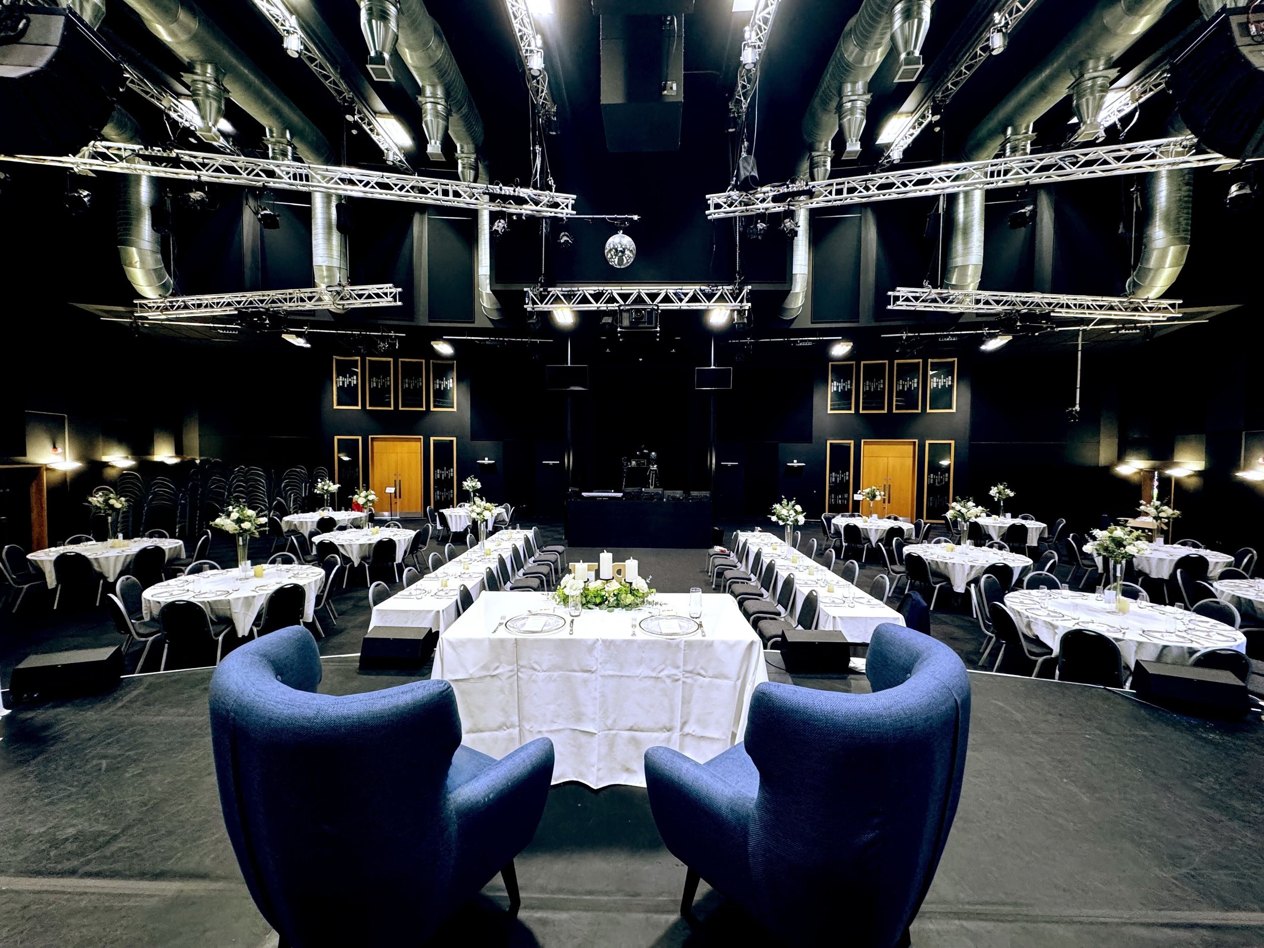A large event space with round tables covered in white tablecloths, decorated with flowers, and black chairs, set for dining. The room has stage lighting, audio equipment, and a disco ball hanging from the ceiling.