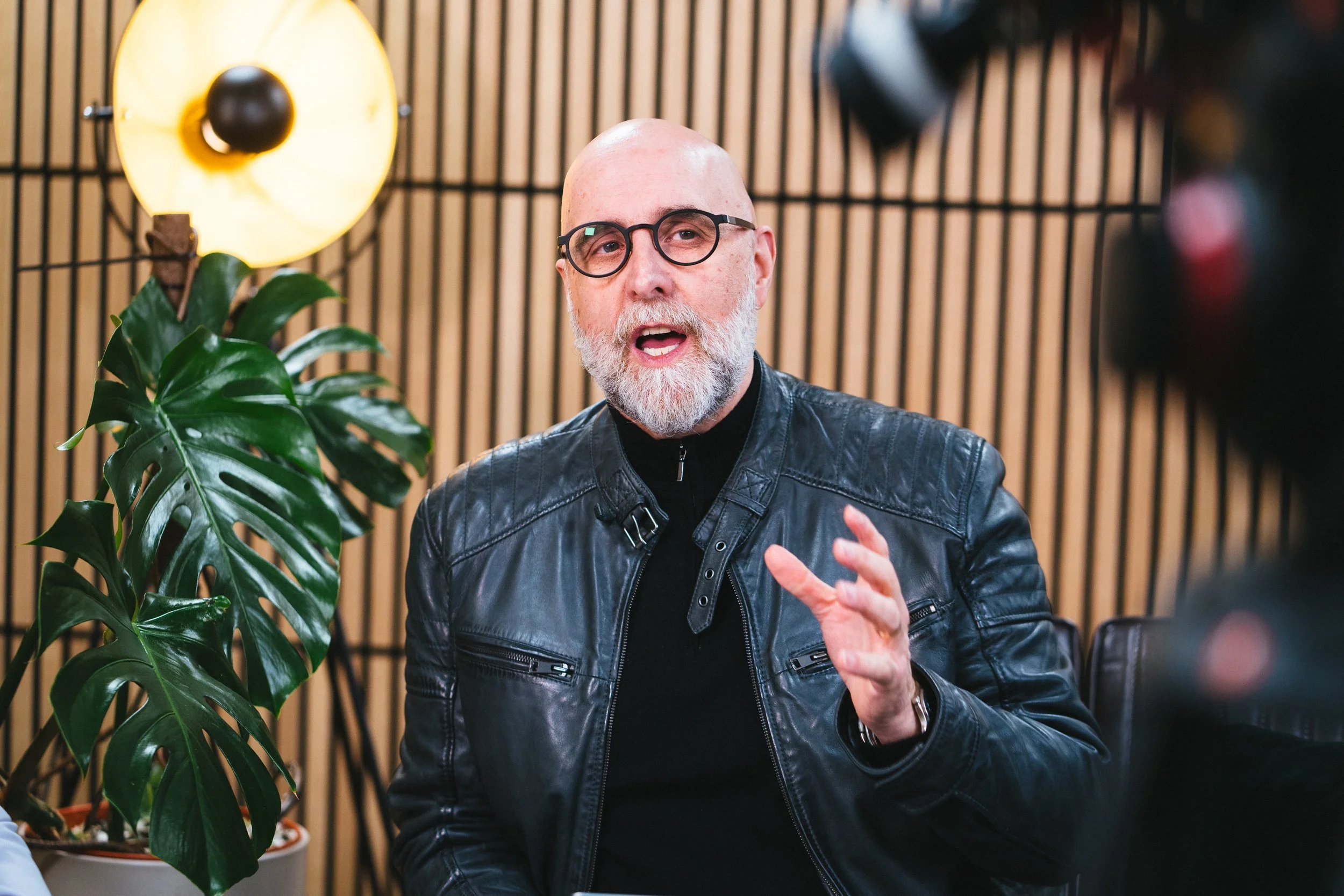 A bald, bearded man wearing glasses and a black leather jacket gesturing while speaking, seated in front of a wooden slat wall with a green leafy plant nearby.