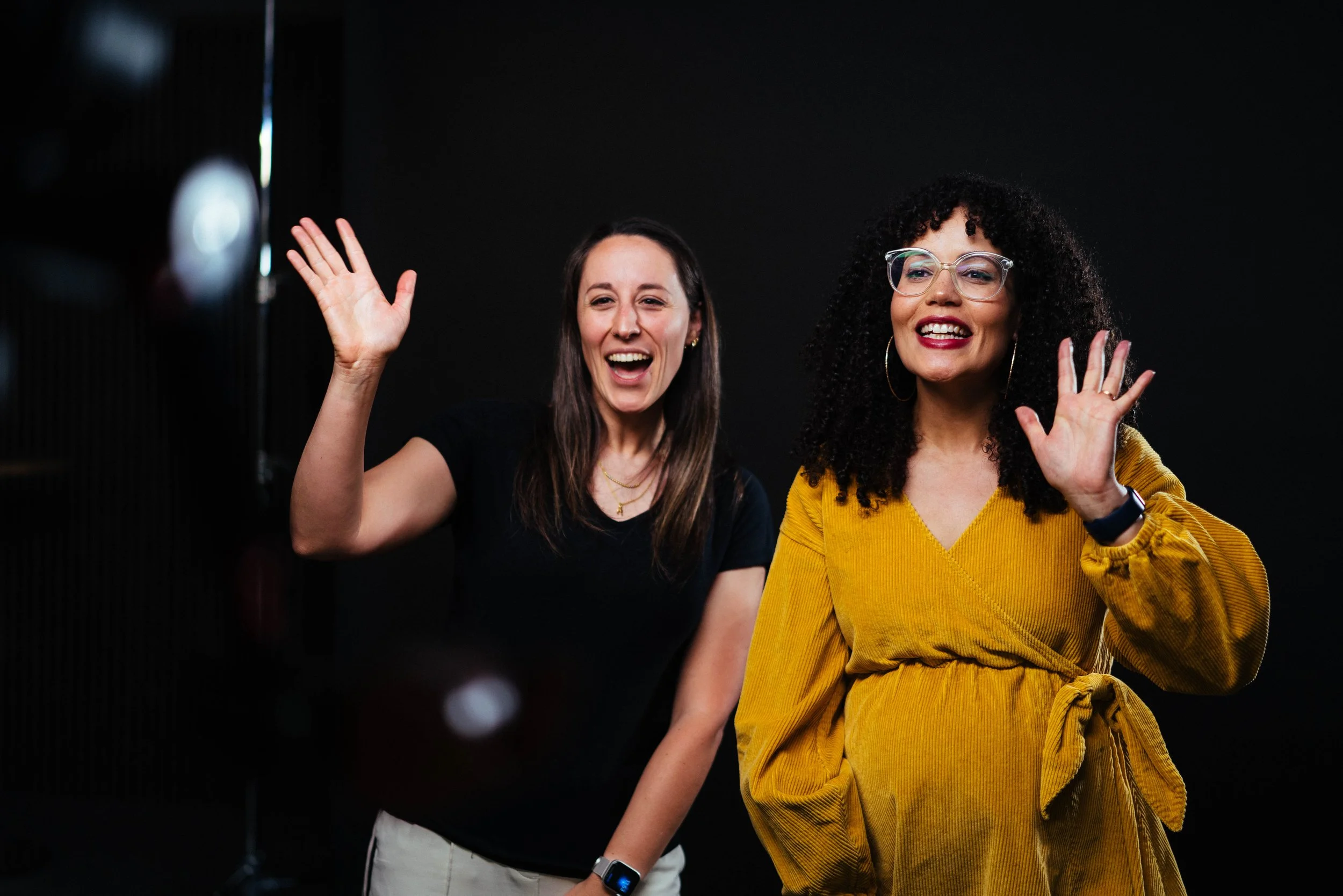 Two women smiling and waving at the camera against a black background.