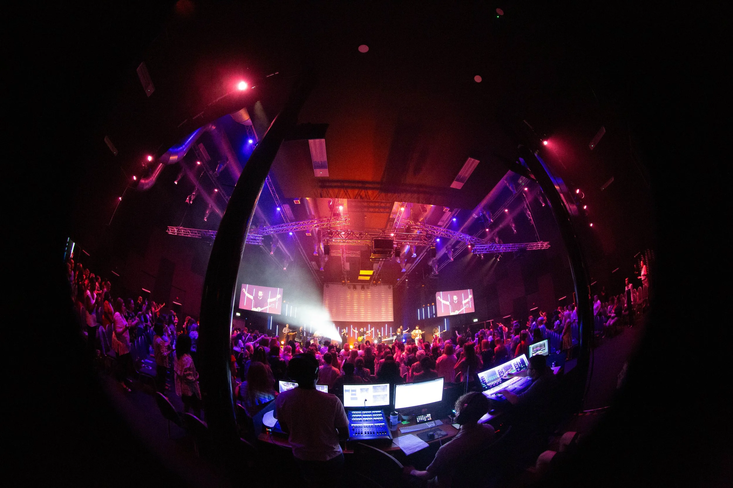 Concert hall filled with audience, stage with performers, colorful lighting, and screens showing live visuals.