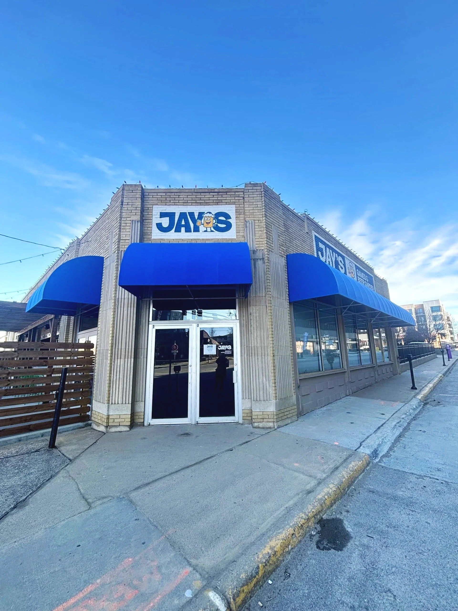 Front view of a building with a sign that reads 'JAY'S' and features a cartoon beer mug. The building has large windows and blue awnings, situated on a city sidewalk with a curb and street in the foreground.