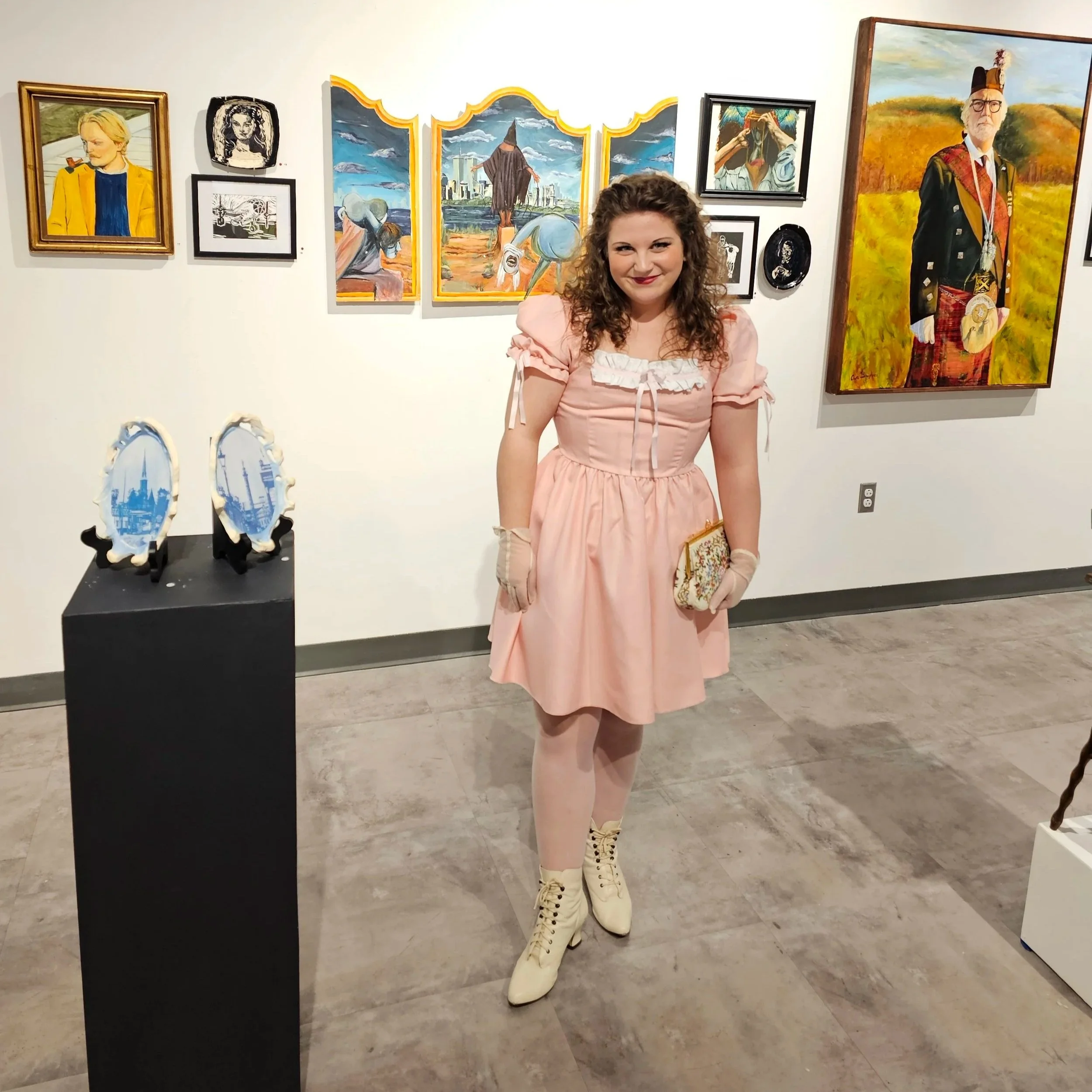 A young woman with long curly hair wearing cream-colored lace-up boots and a pink dress standing in an art gallery, smiling at the camera. Behind her, various colorful paintings, including a large portrait of a man in traditional Scottish attire and several landscape scenes, are displayed on the wall. To the left, there is a black pedestal with decorative items that appear to be miniature replicas of famous landmarks.