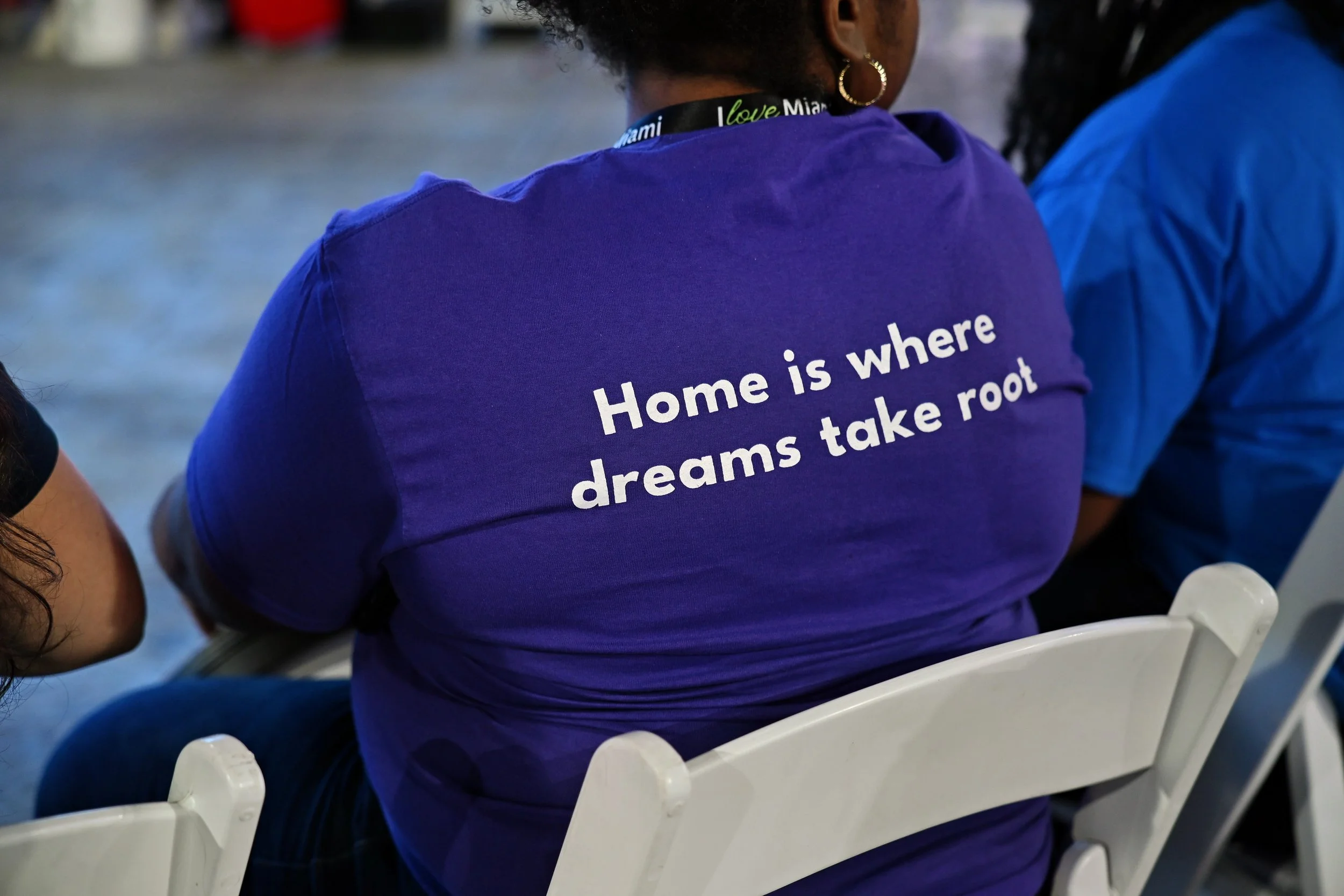 Back view of a woman wearing a purple t-shirt with the quote 'Home is where dreams take root' printed on the back, sitting on a white chair at an outdoor event.