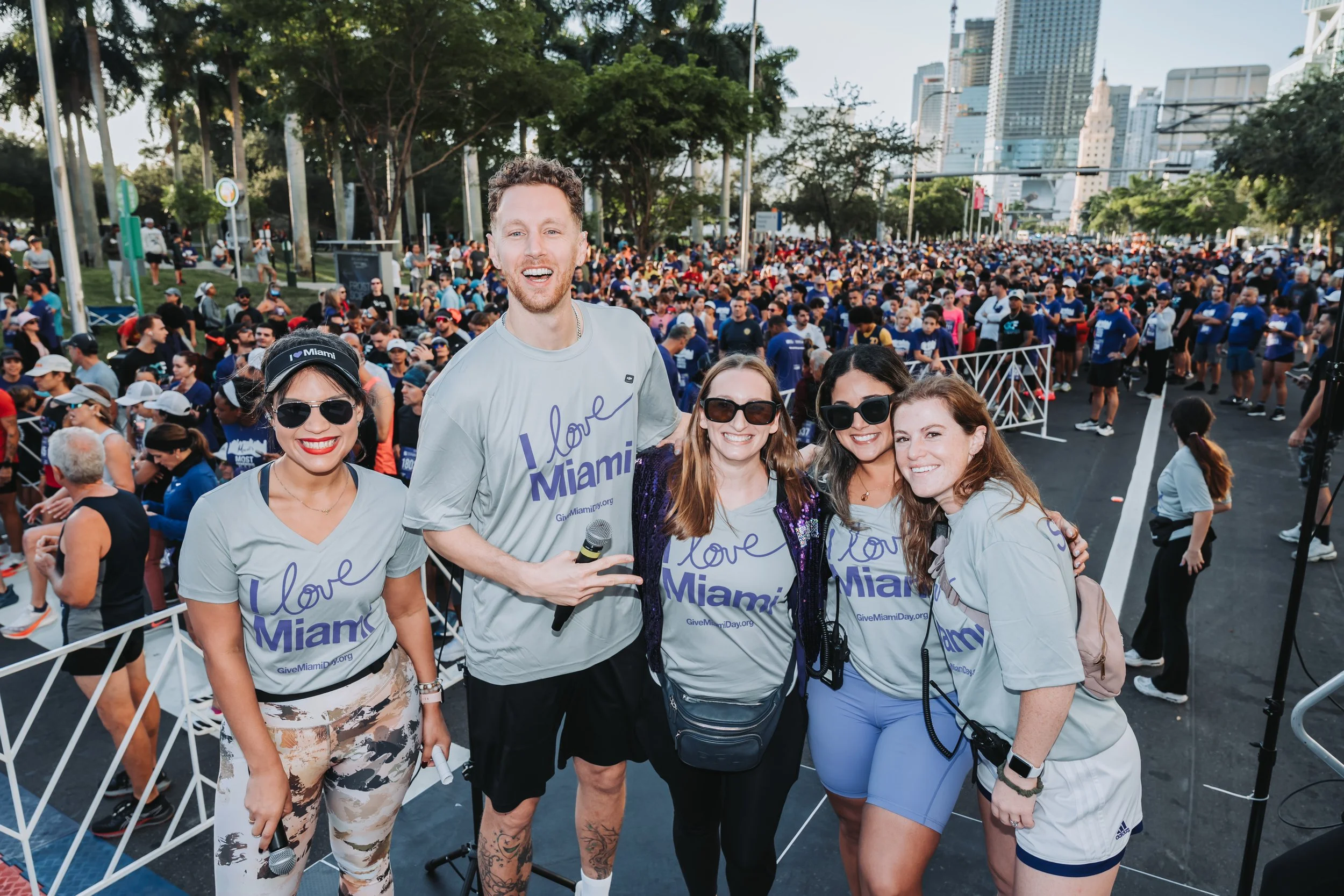 Group of five smiling people at an outdoor event, wearing matching 'I love Miami' t-shirts, with a large crowd and city skyline in the background.