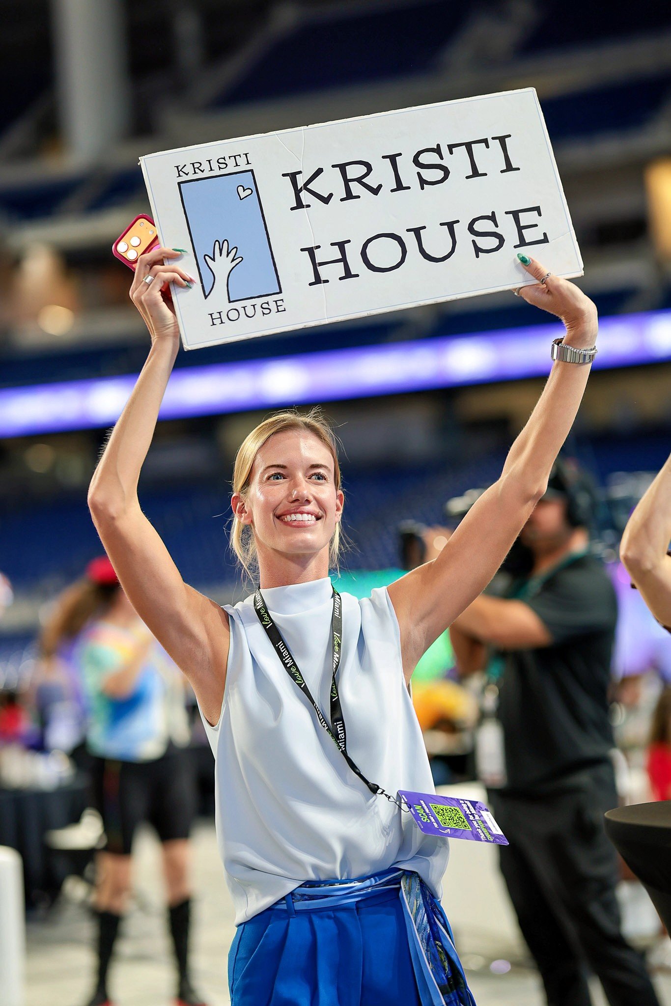 A woman smiling and holding a sign that reads "Kristi House" during an indoor event. She is wearing a sleeveless top, blue shorts, and has a lanyard with a badge around her neck.