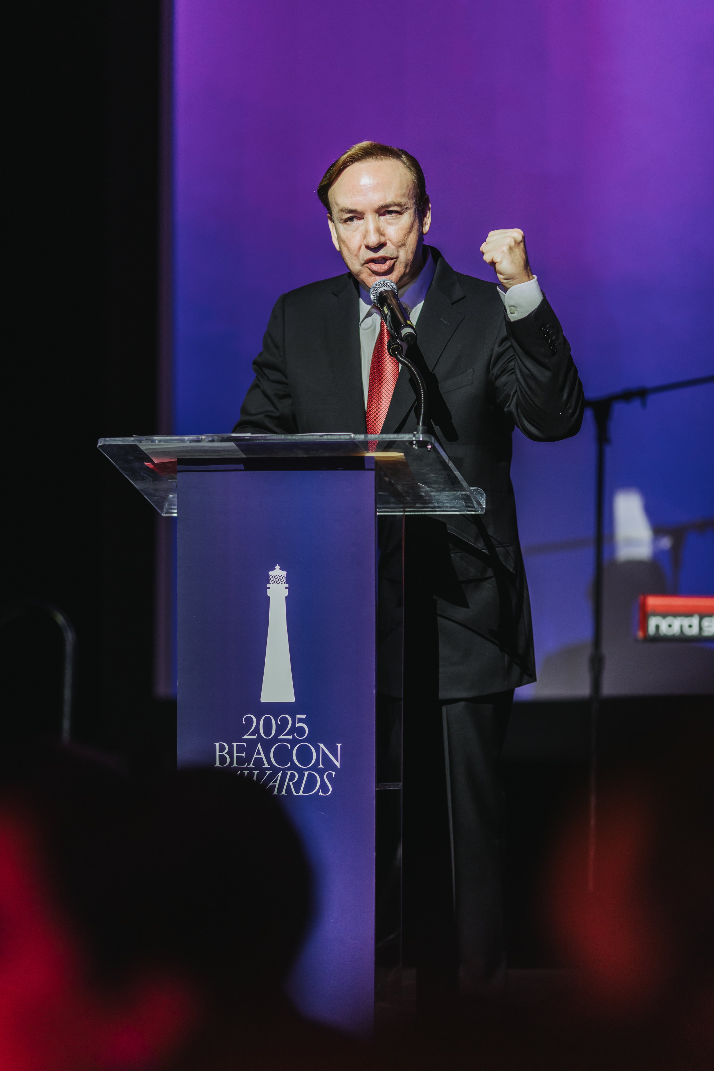 A man in a black suit and red tie speaking at a podium during the 2025 Beacon Awards, with a purple background. The podium has a lighthouse logo and text reading '2025 BEACON AWARDS'.