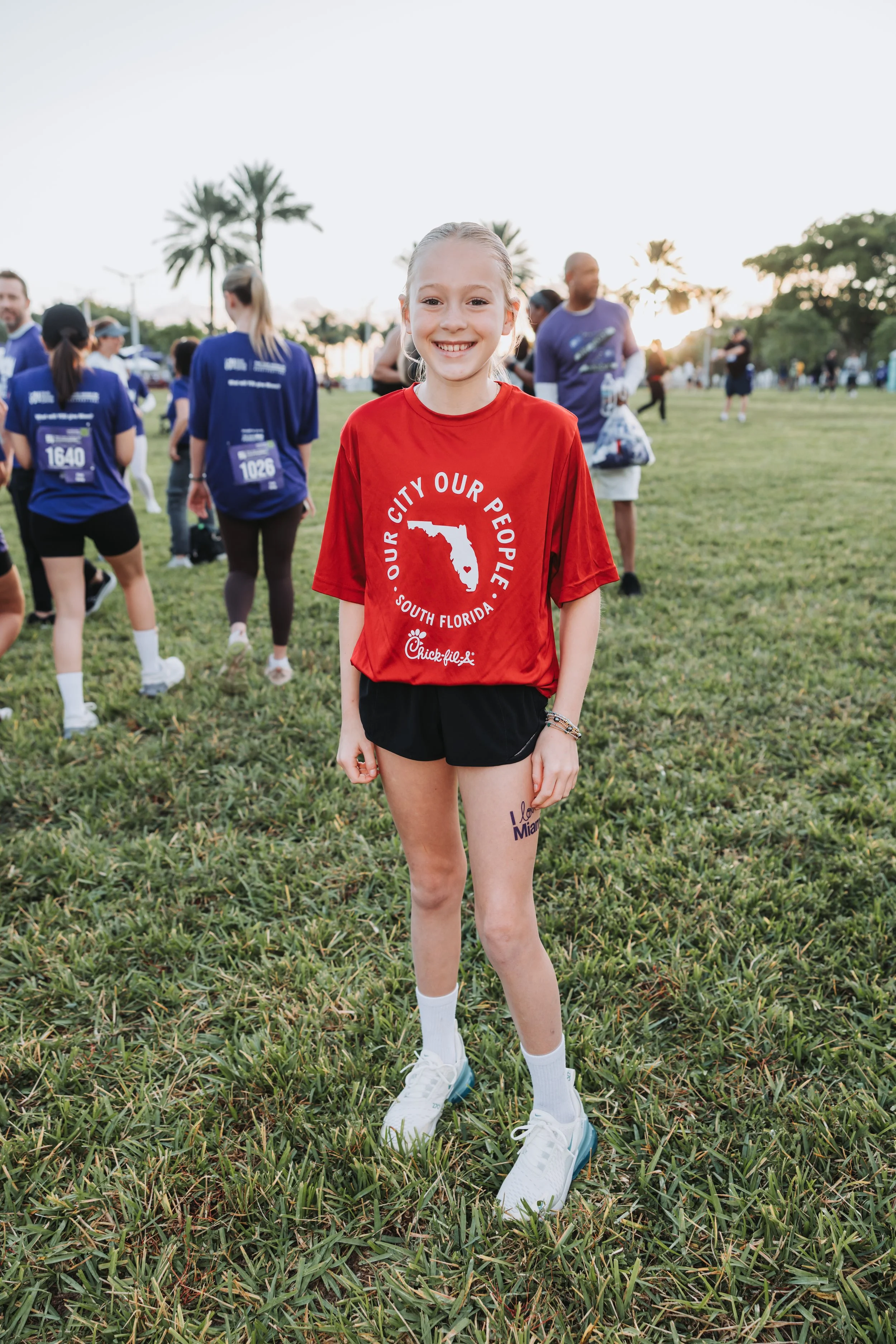 A young girl with blonde hair smiling at the camera during a race event at a park in Florida, wearing a red shirt, black shorts, white socks, and sneakers, with people in numbered shirts in the background.