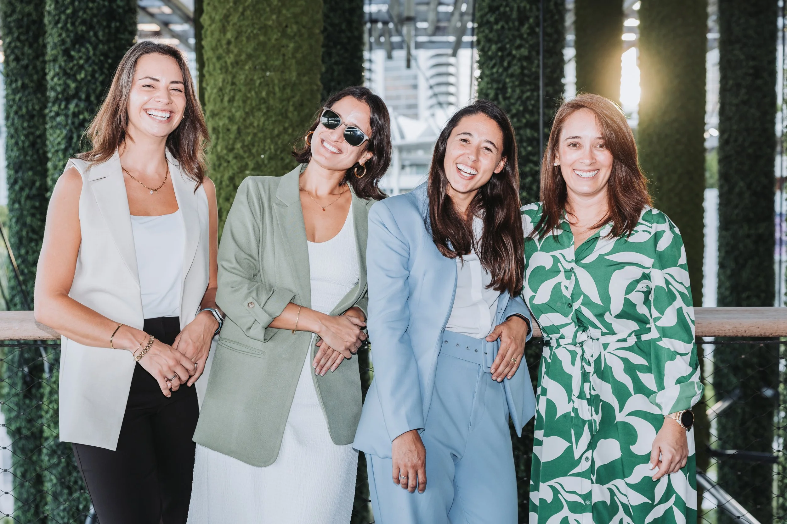 Group of four women smiling and standing together outdoors in front of green plants and buildings.