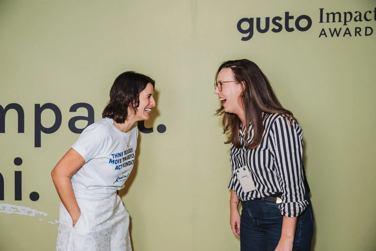 Two women are smiling and laughing while facing each other at an event with a green backdrop that has the words 'gusto Impact Award' on it.