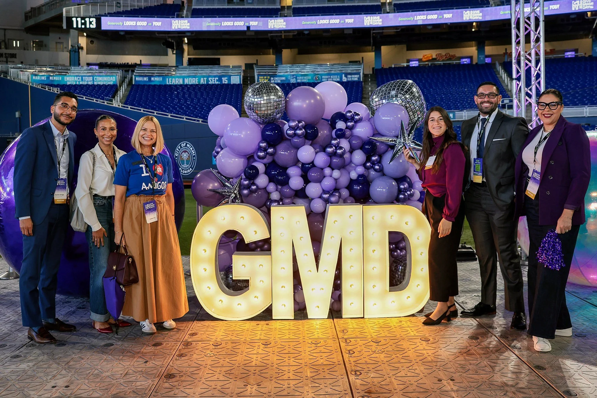 Group of eight diverse people standing in front of an illuminated sign that reads GMD, with purple and silver balloons and decorative stars, inside a stadium or large indoor venue.