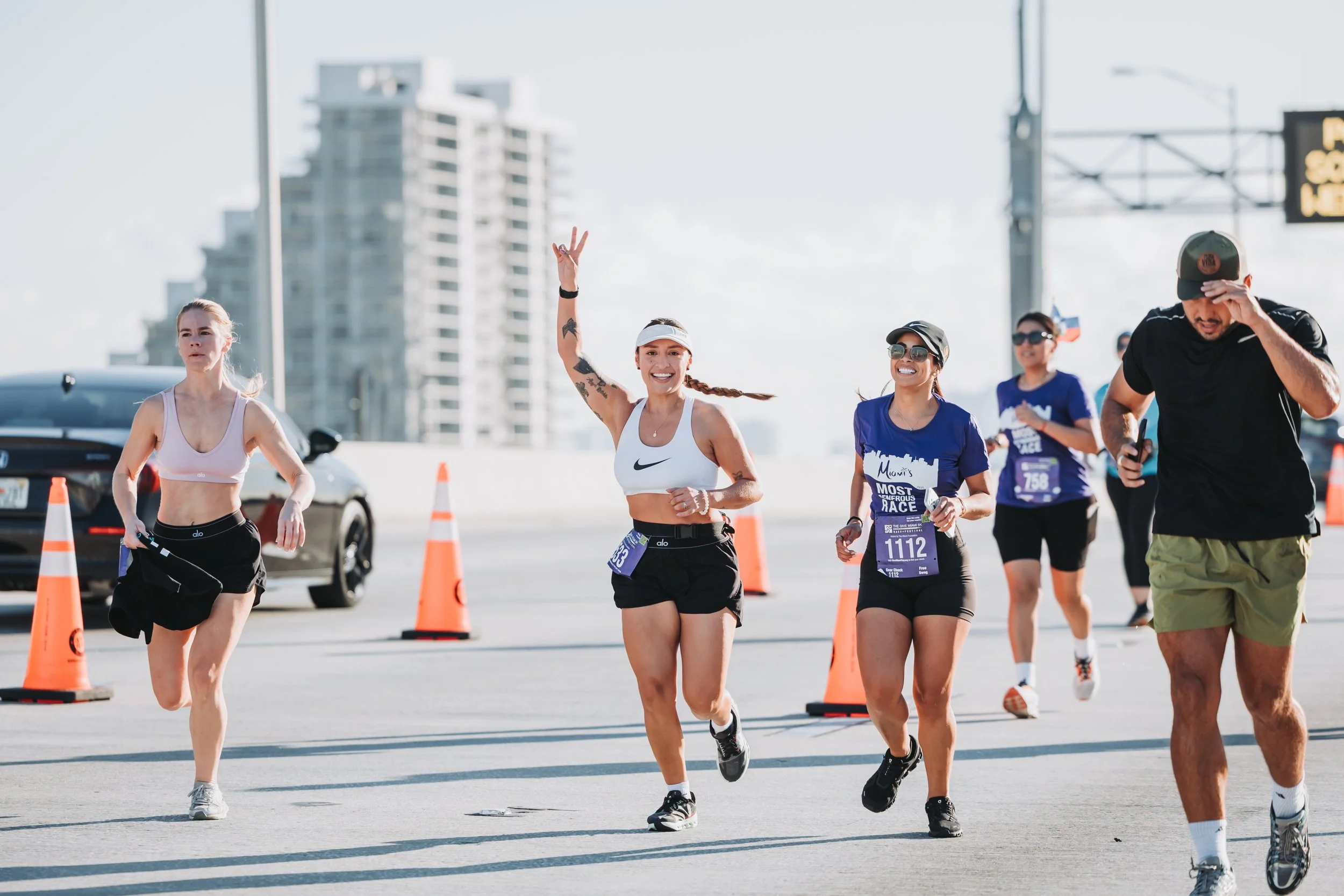 Group of runners participating in a marathon, running on a city street with orange cones and high-rise buildings in the background.