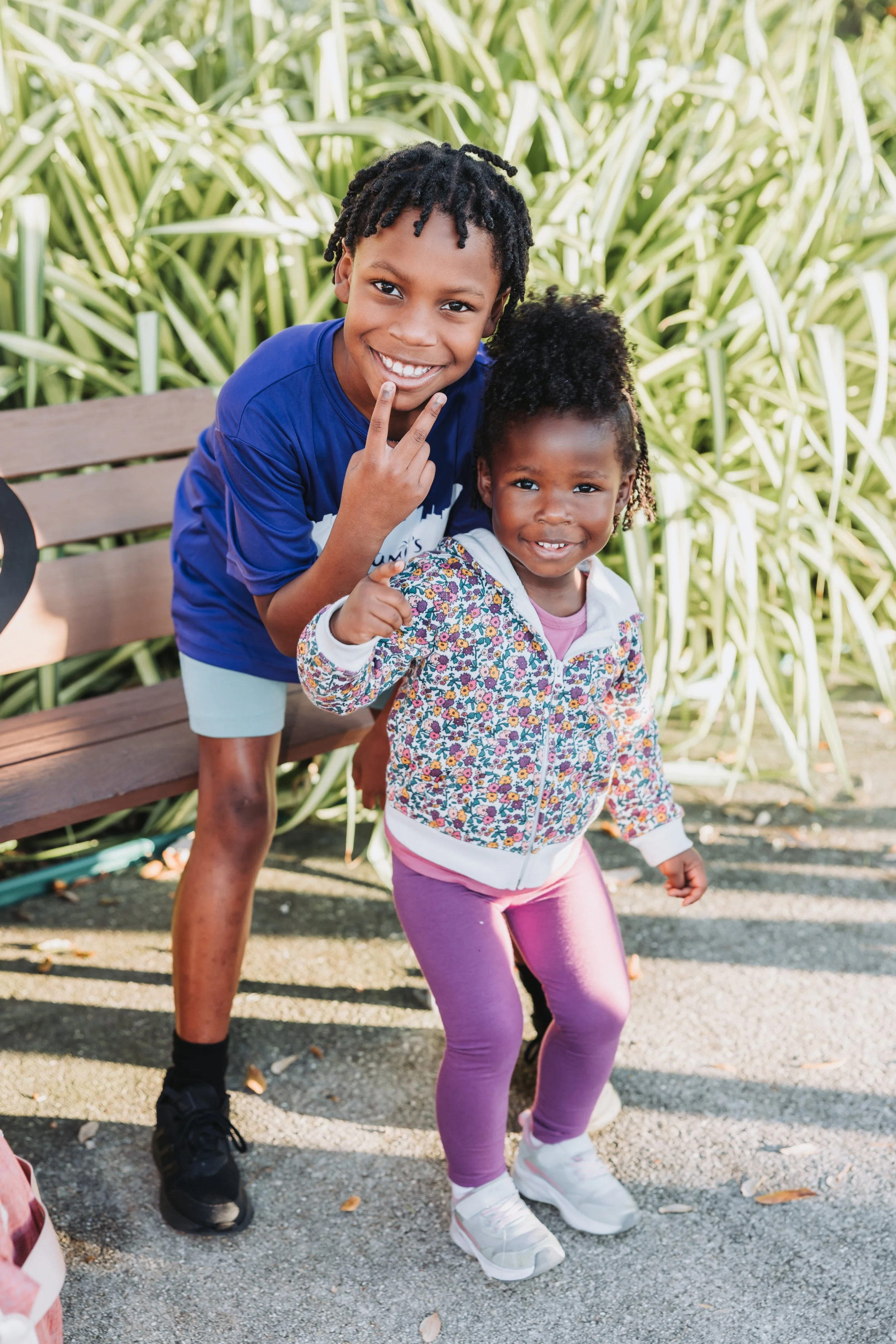 Two children, a boy and a girl, smiling outdoors. The boy is making a peace sign with his fingers and leaning over a bench, while the girl is standing in front, dressed in purple leggings and a floral jacket, smiling at the camera.