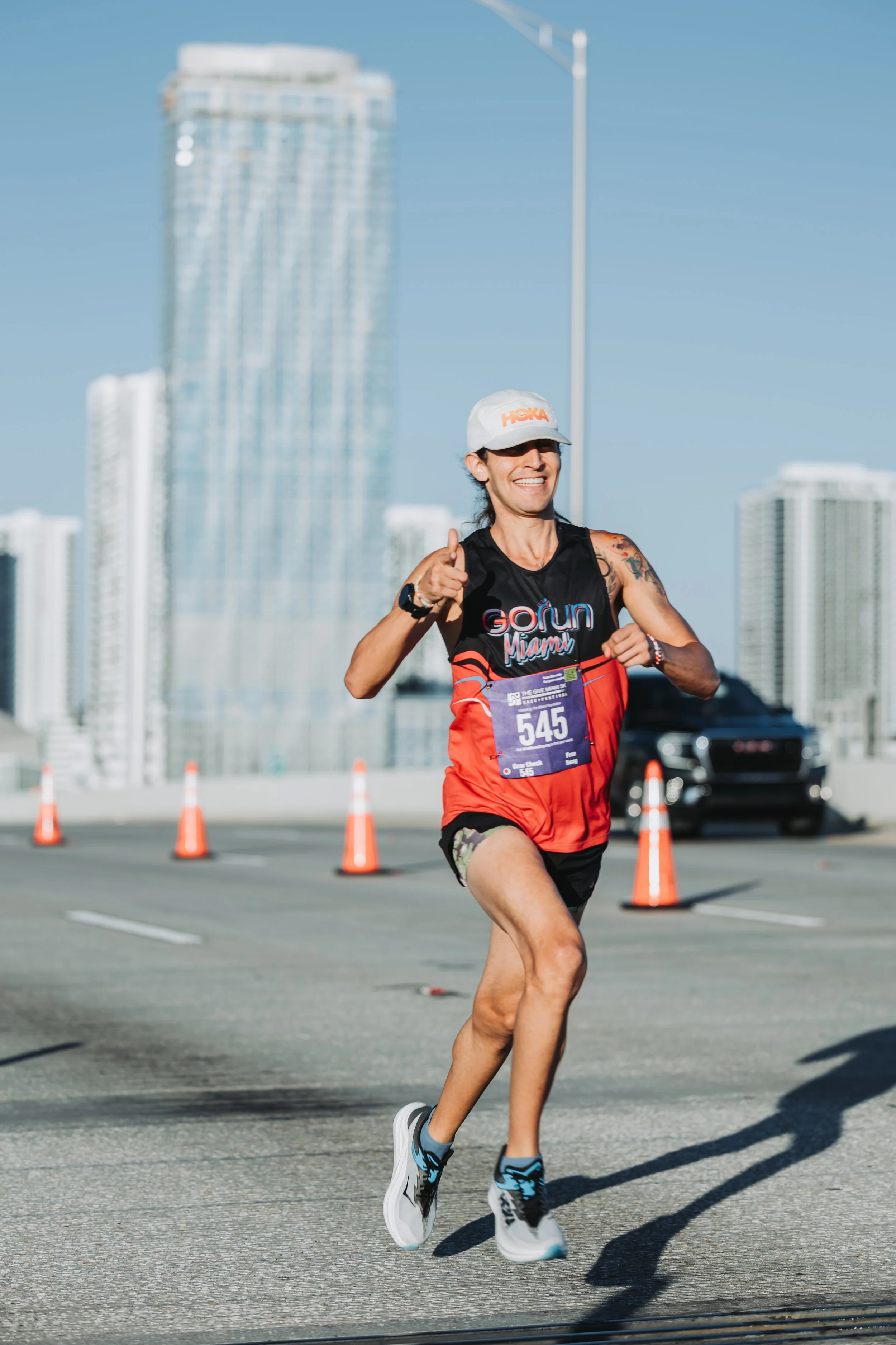 A woman running in a marathon, smiling, wearing athletic gear, a white cap, and a race bib number 545, with city skyscrapers and orange traffic cones in the background.