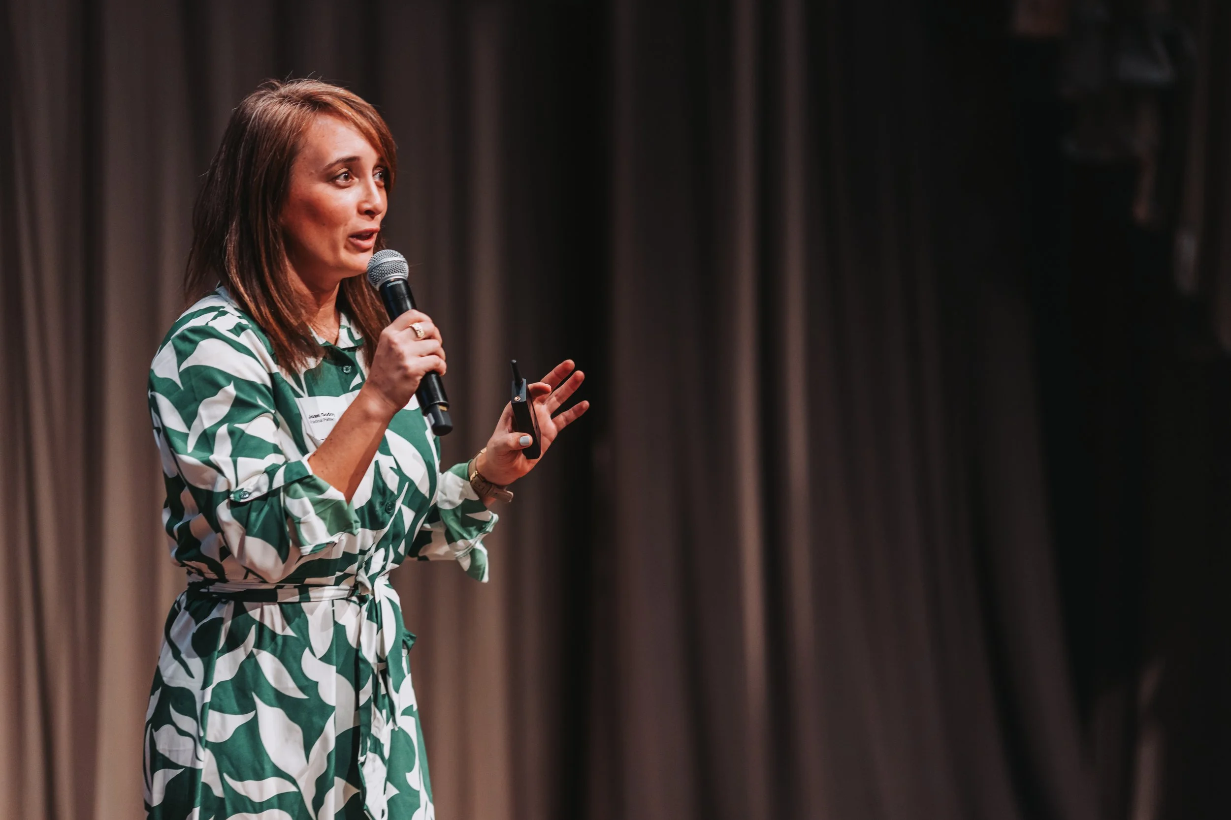 A woman with shoulder-length brown hair, wearing a green and white patterned dress, holding a microphone and speaking on stage with a brown curtain backdrop.