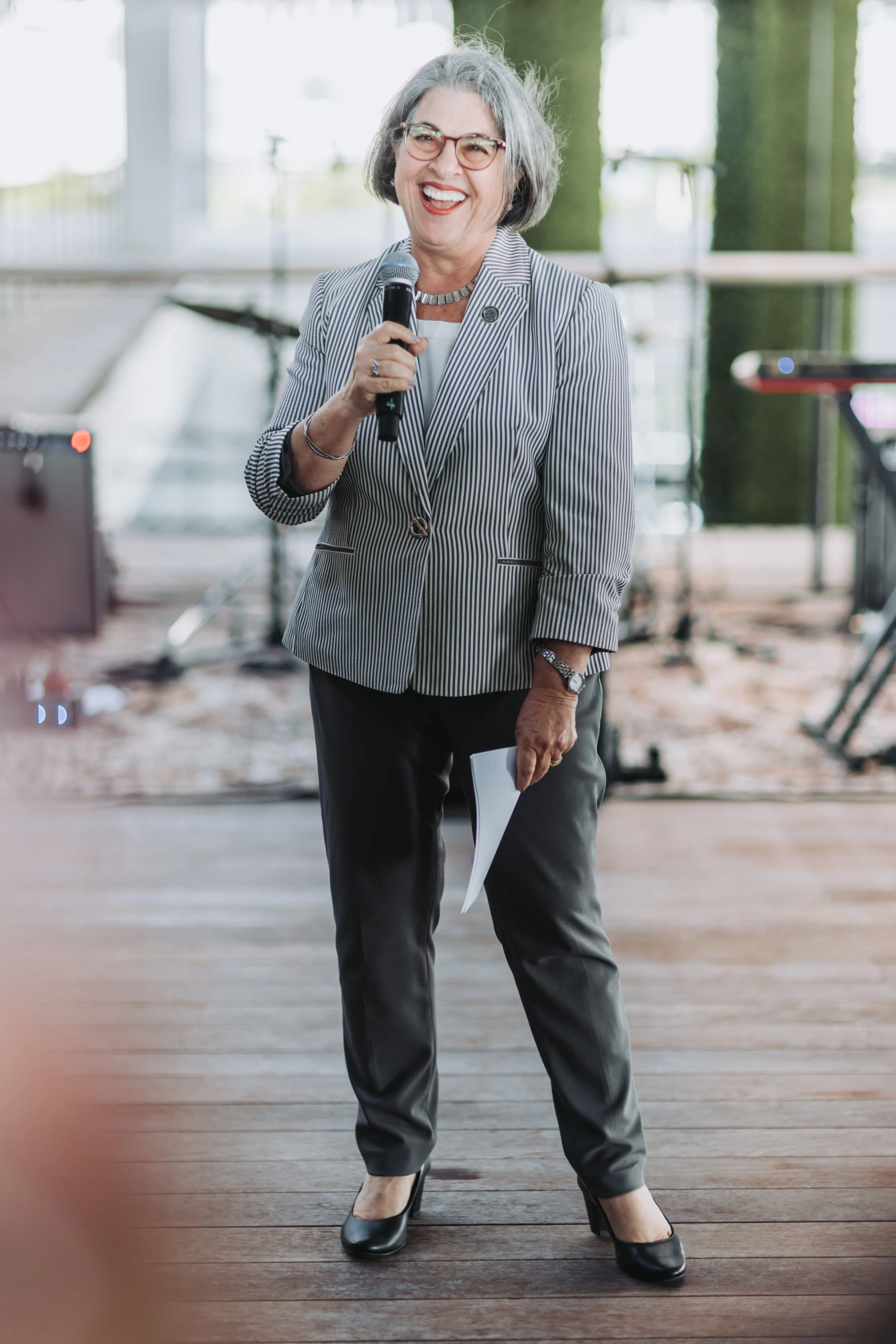 A smiling older woman with gray hair and glasses, dressed in a striped blazer and dark pants, holding a microphone and a piece of paper, speaking at an indoor event.