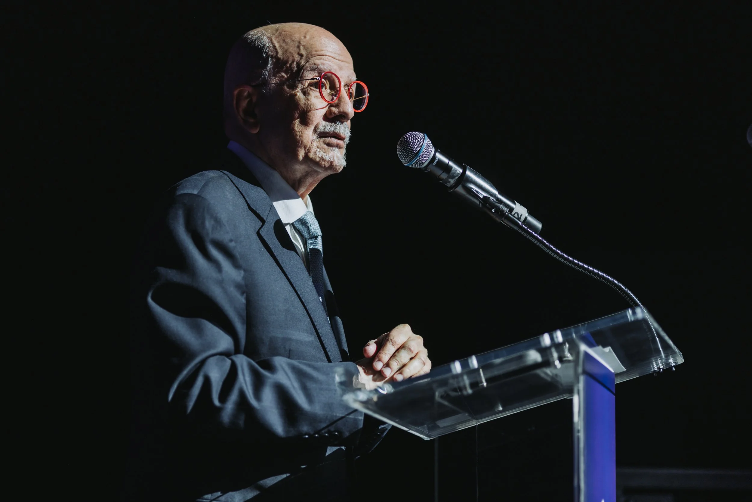 An elderly man with glasses and a bald head, wearing a dark suit, stands at a clear podium speaking into a microphone against a dark background.