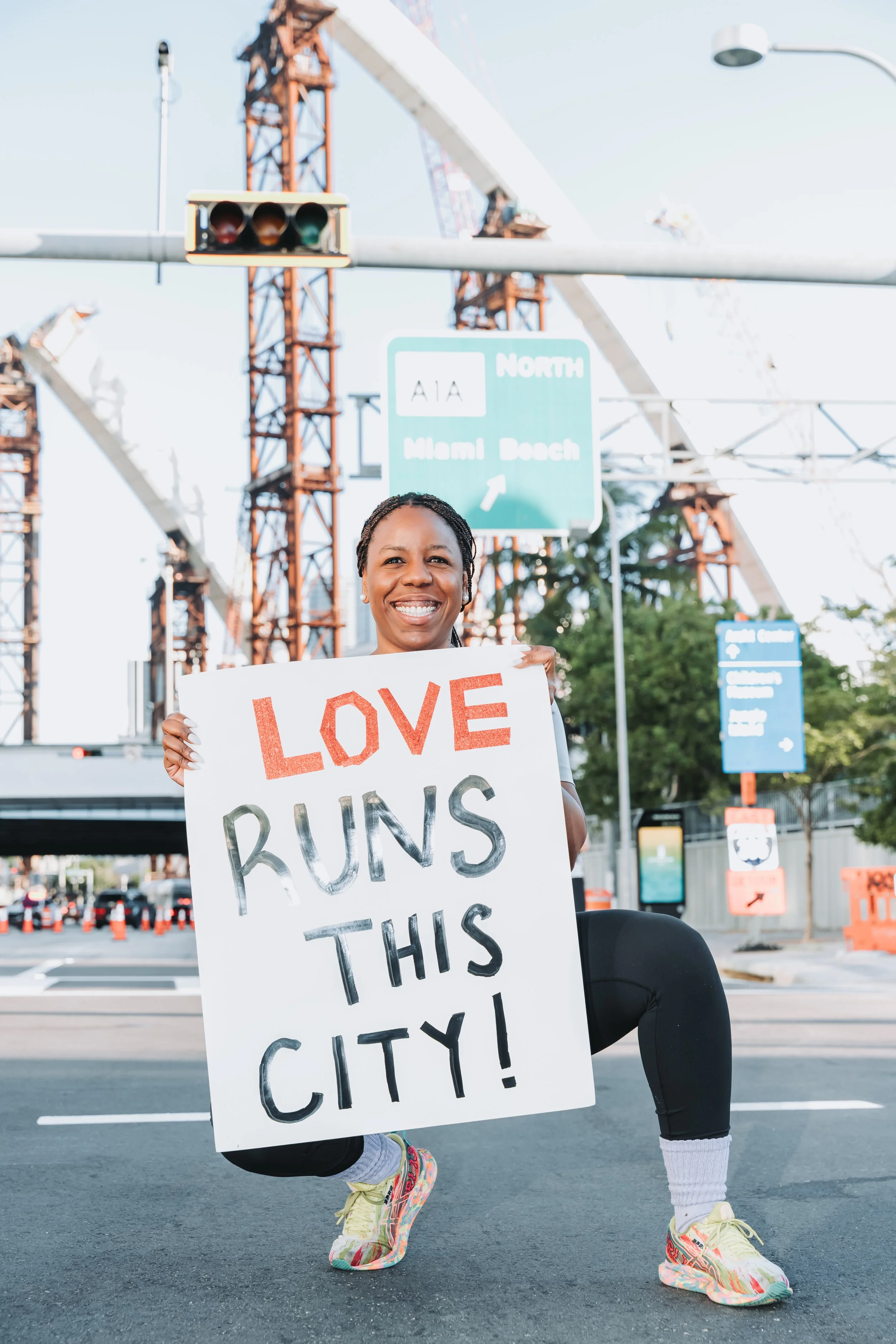 Woman smiling and kneeling on city street holding a sign that says "Love Runs This City!" with construction cranes and traffic signs in the background