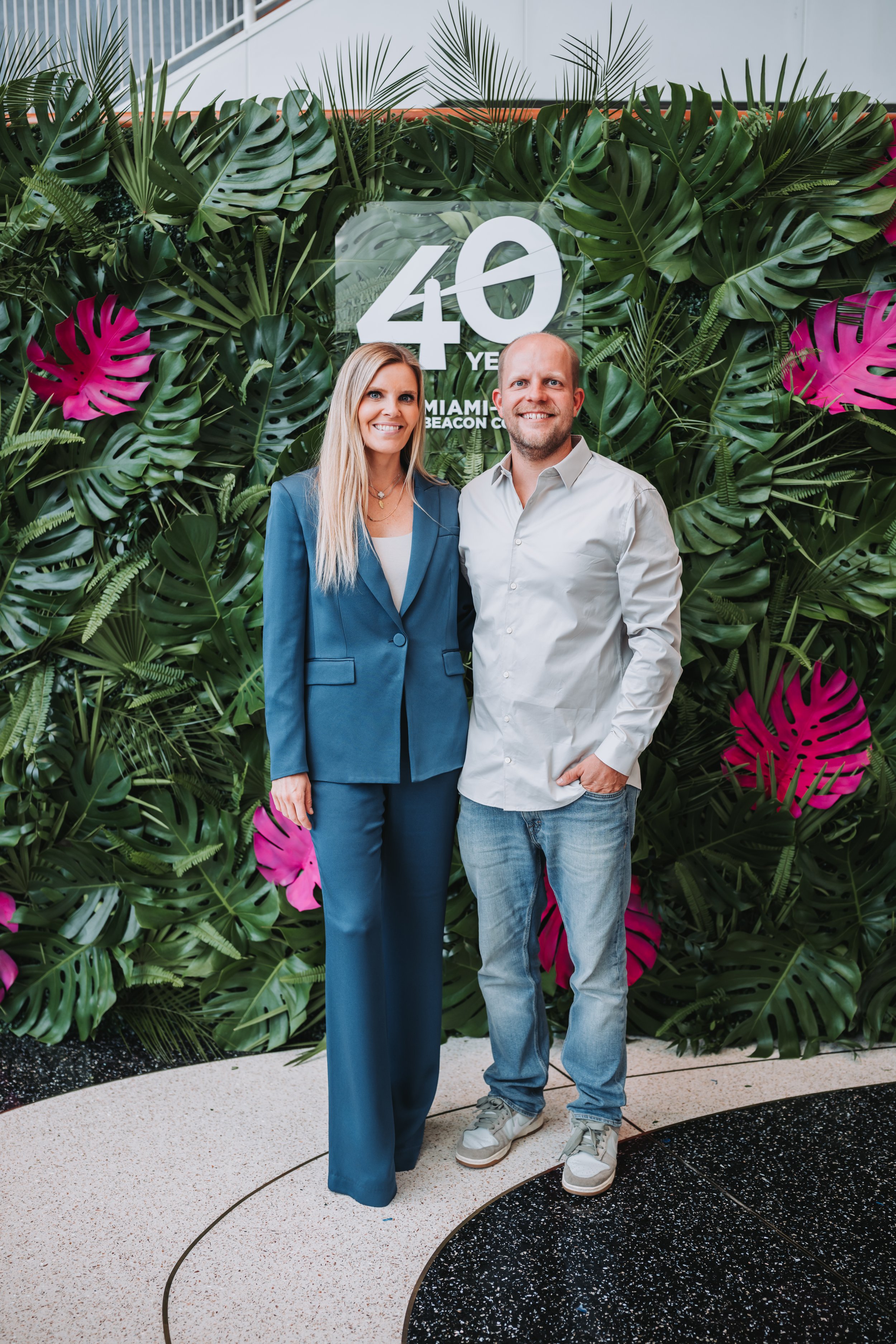 A man and woman standing in front of a tropical plant backdrop with a sign that reads '40 Years Miami Beach Convention Center'