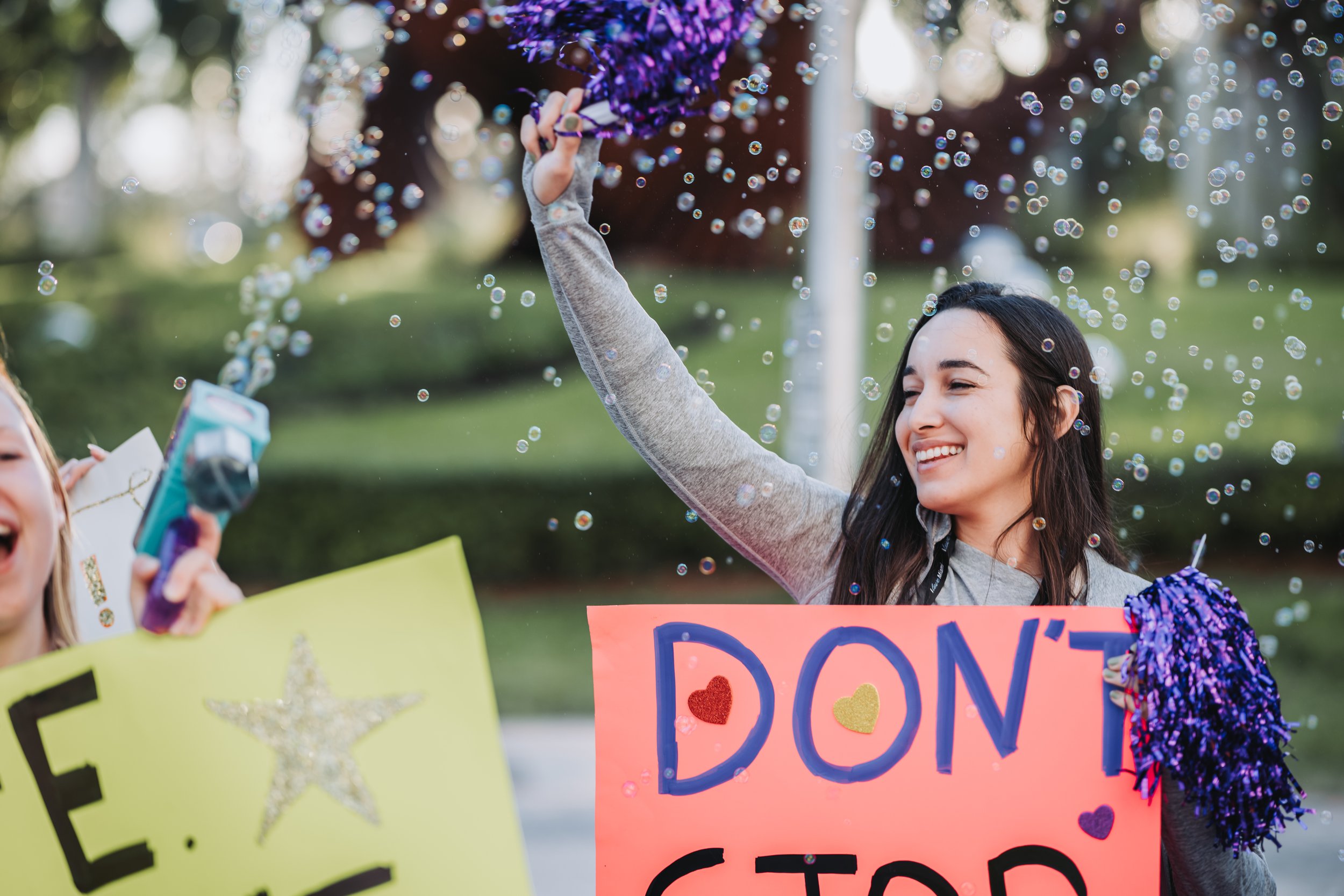 Smiling young woman holding a purple pom-pom in one hand and a sign that says 'DON'T STOP' in colorful letters, surrounded by bubbles and outdoors during daytime.