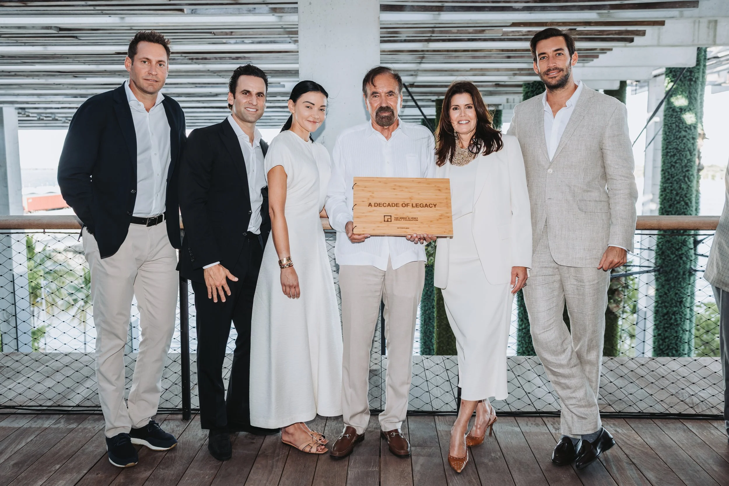 Jorge M. Pérez family standing on a wooden deck, with the man in the center holding a plaque that reads "A DECADE OF LEGACY".