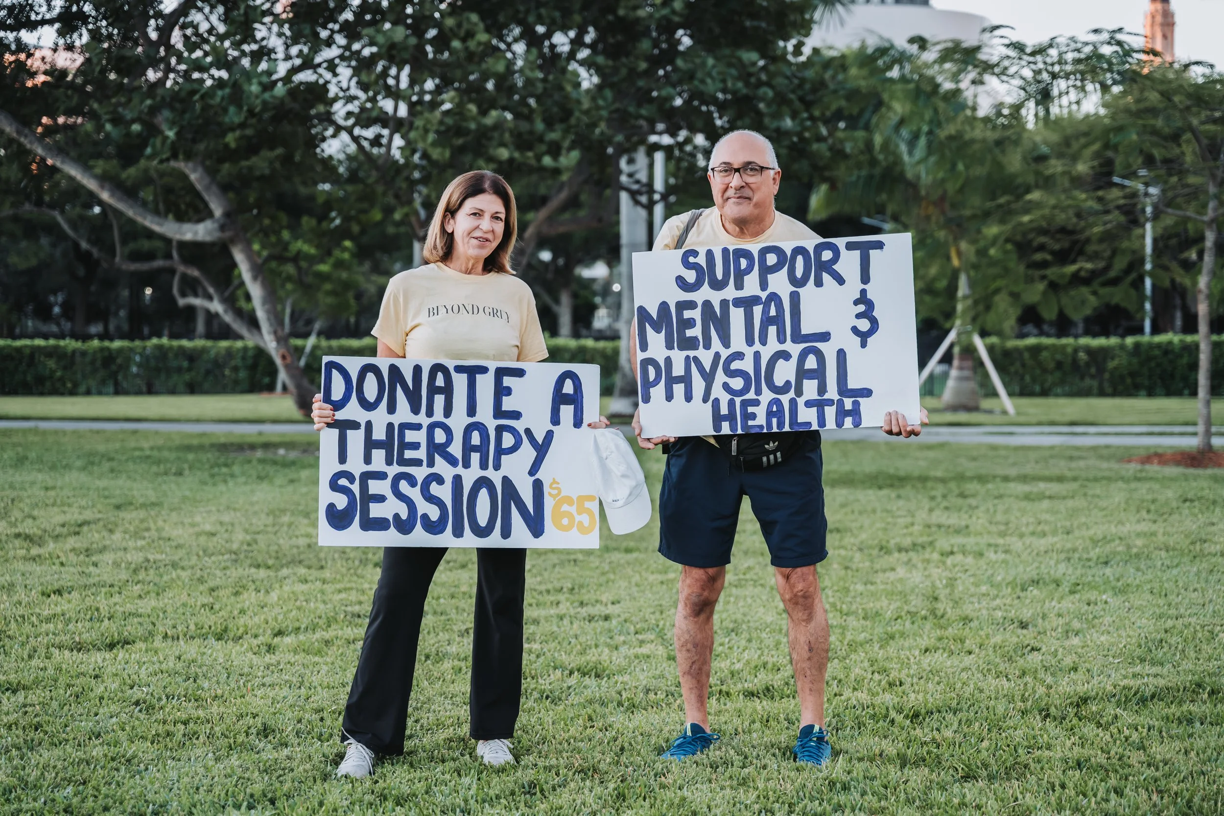 A woman and a man stand on grass in a park holding signs advocating for healthcare. The woman's sign reads "Donate a Therapy Session $65" and the man's sign says "Support Mental & Physical Health." They are smiling and dressed casually.