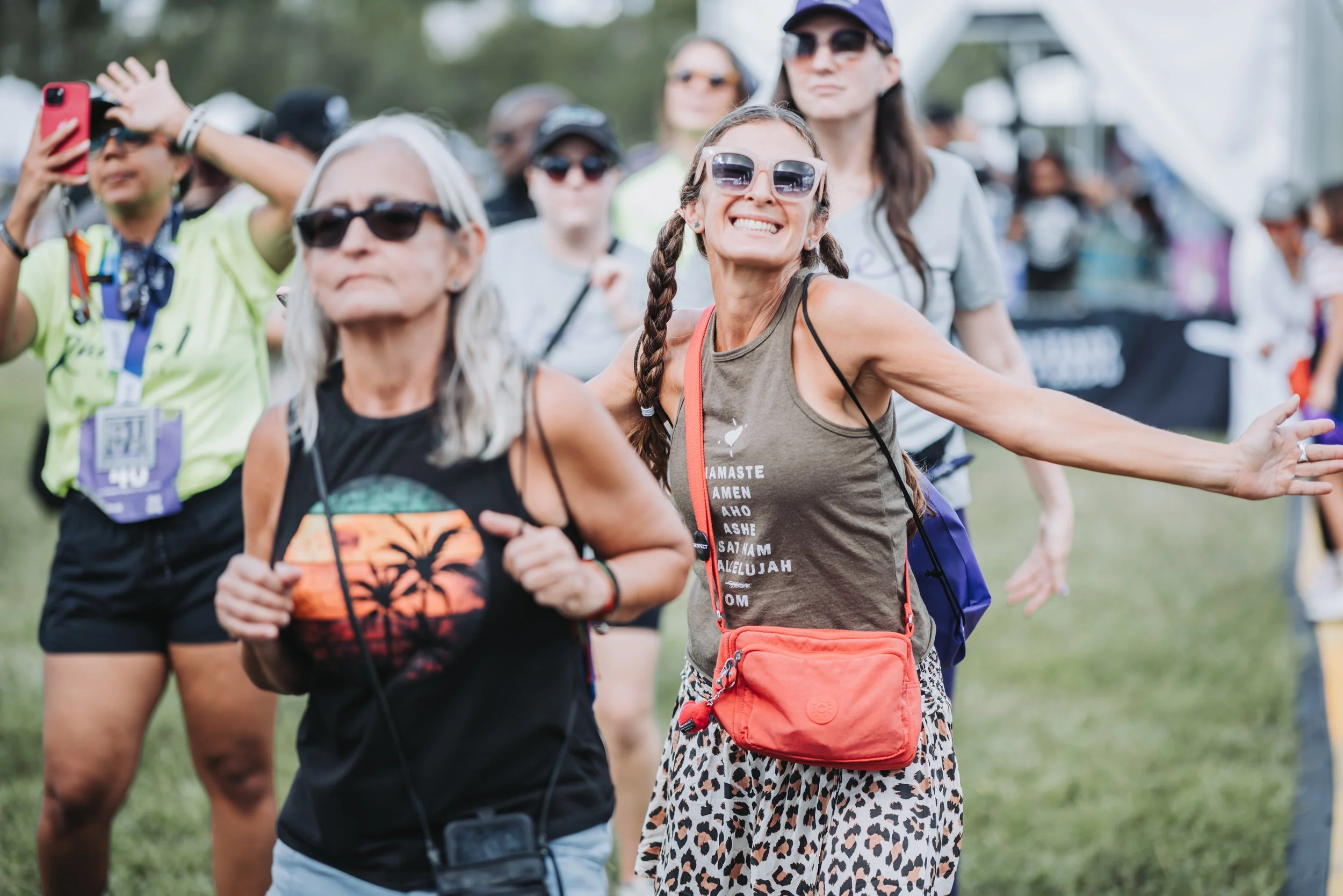 Group of women participating in an outdoor event or race, some smiling, wearing casual athletic clothing, sunglasses, and carrying small bags, with grassy field and tents in the background.