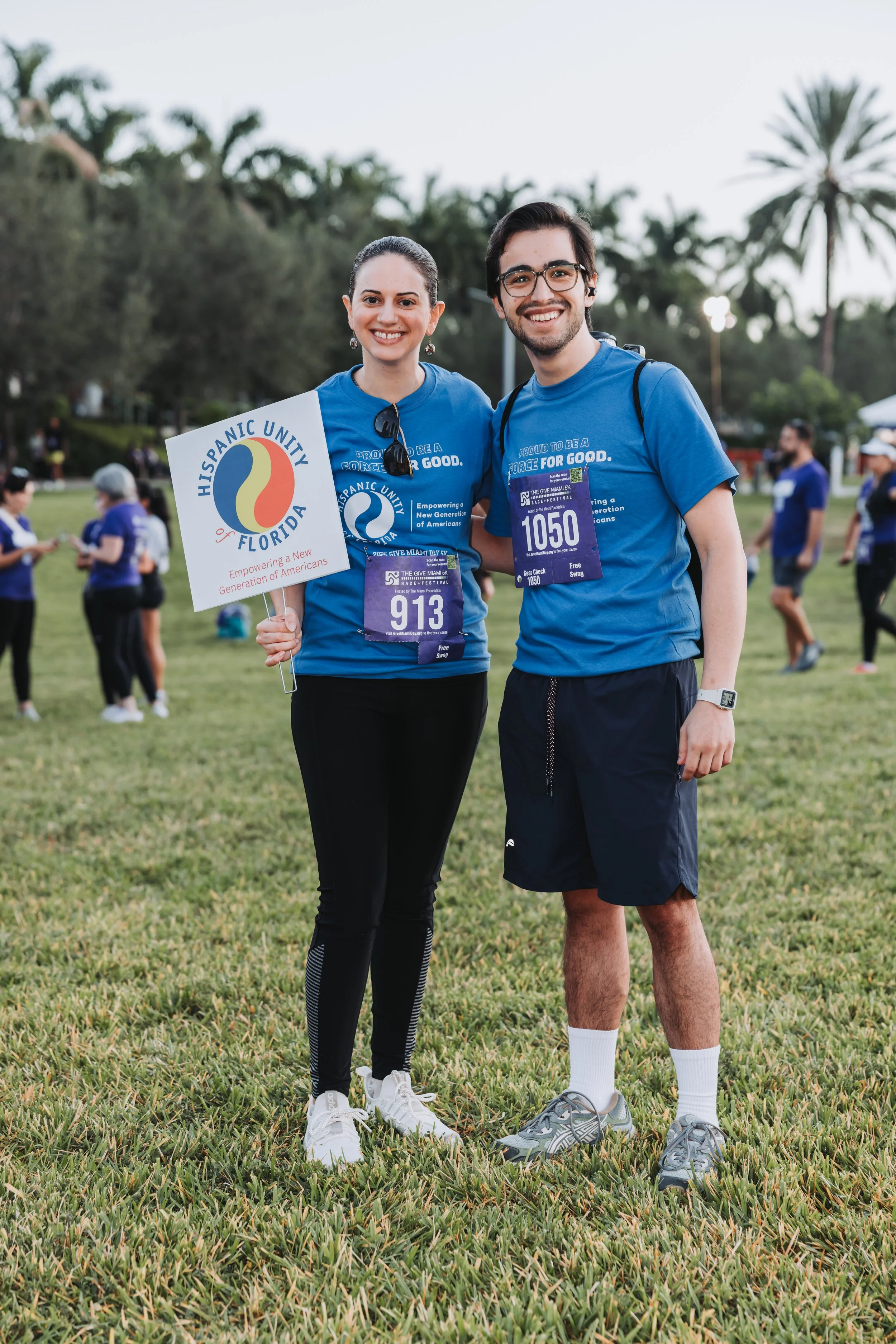 Two people standing on grass at an outdoor event, smiling at the camera. They are wearing blue T-shirts with event logos, athletic shorts/pants, and sneakers. The woman holds a sign that says 'Hispanic Unity of Florida' and has a bib number 913, whil
