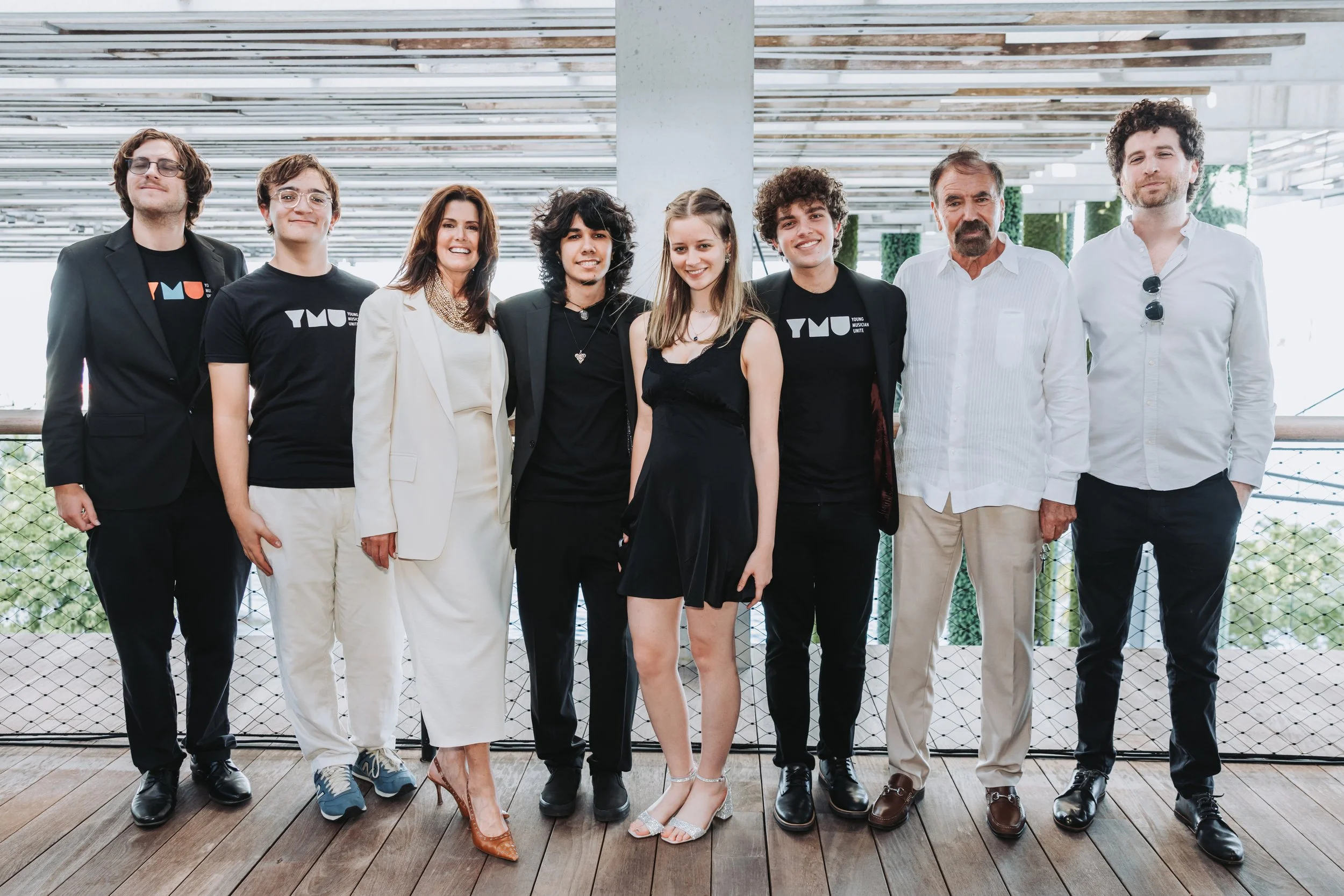 Group photo of nine people standing on a wooden floor under a modern canopy, smiling and dressed in formal to casual attire, with some wearing black T-shirts with a logo.