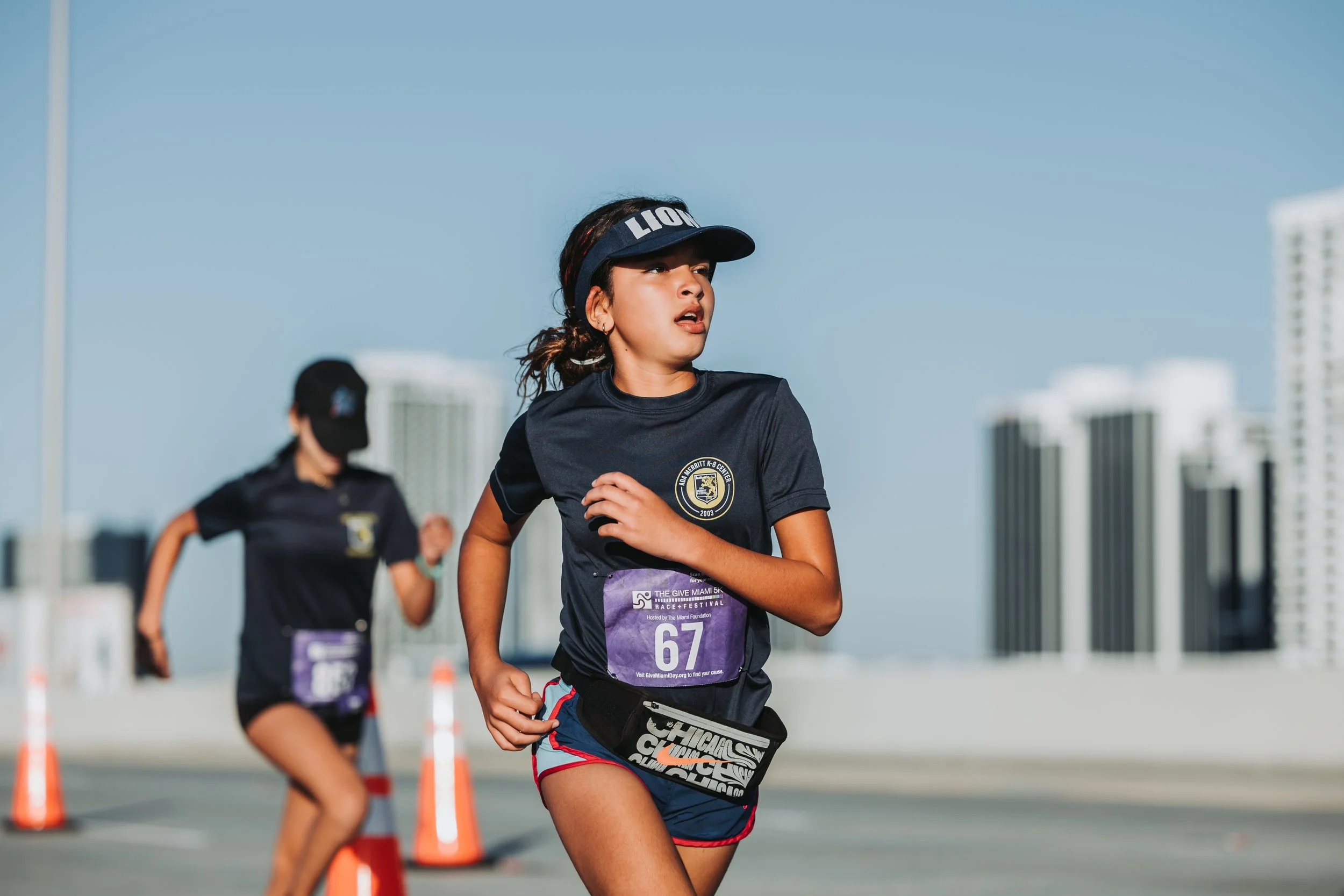 Two young female runners participating in a race on a city street, with tall buildings in the background. One is in focus and the other is slightly blurred.