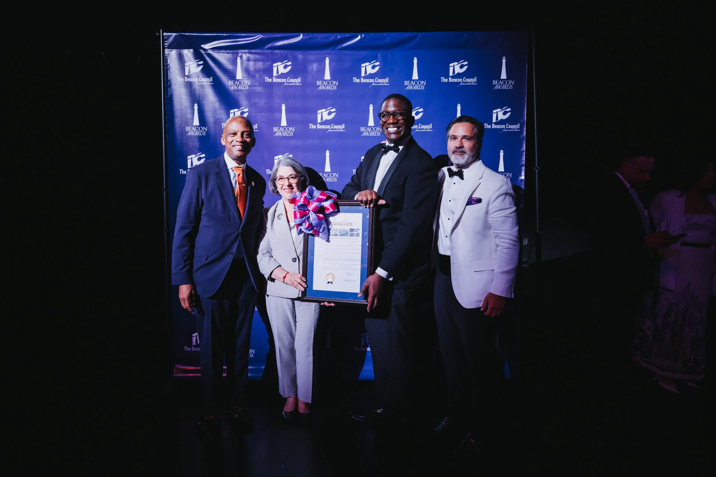 Four people in formal attire standing on stage at an award ceremony, holding a framed certificate with a theater backdrop that reads 'The Beacon Council' and 'BEACON AWARDS'.