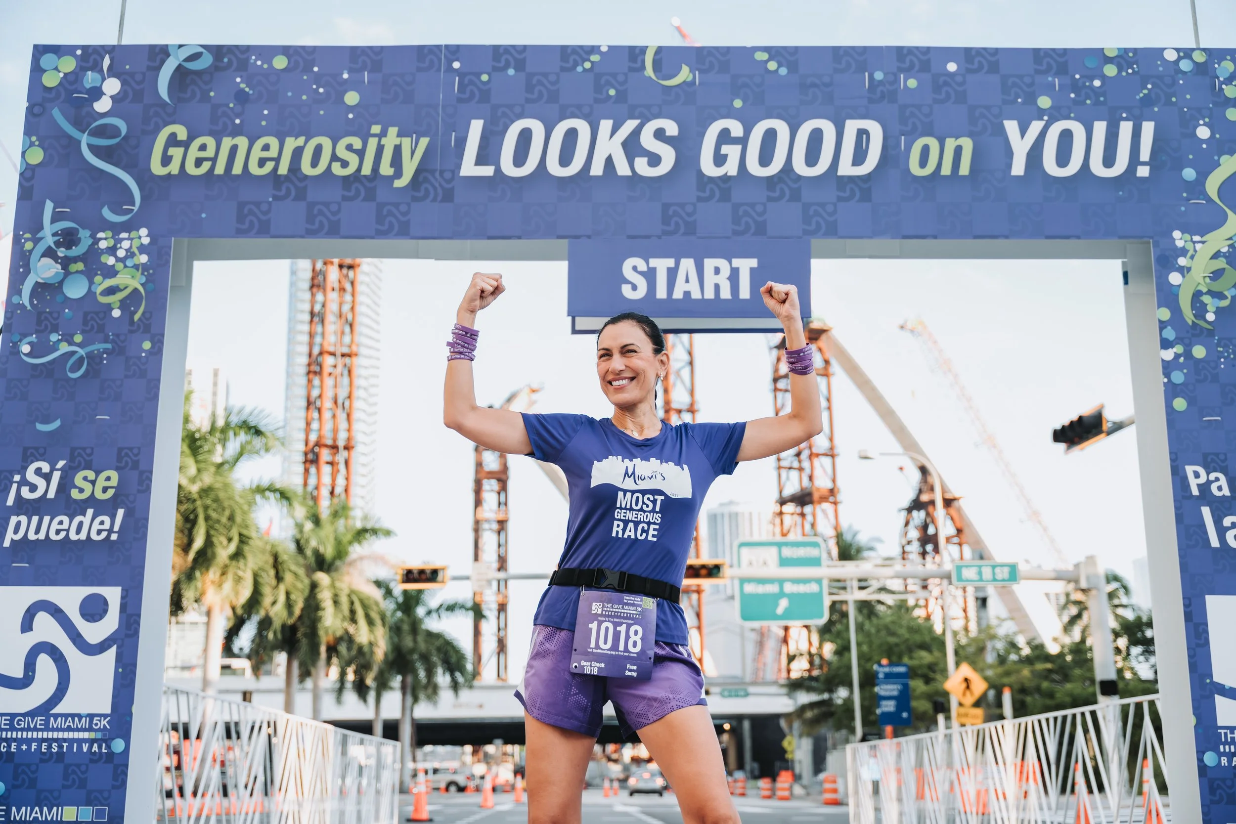 A woman smiling and flexing her arms at the start line of a race event, wearing a blue shirt and purple shorts, with a race bib number 1018, in Miami, Florida, with construction cranes and palm trees in the background.