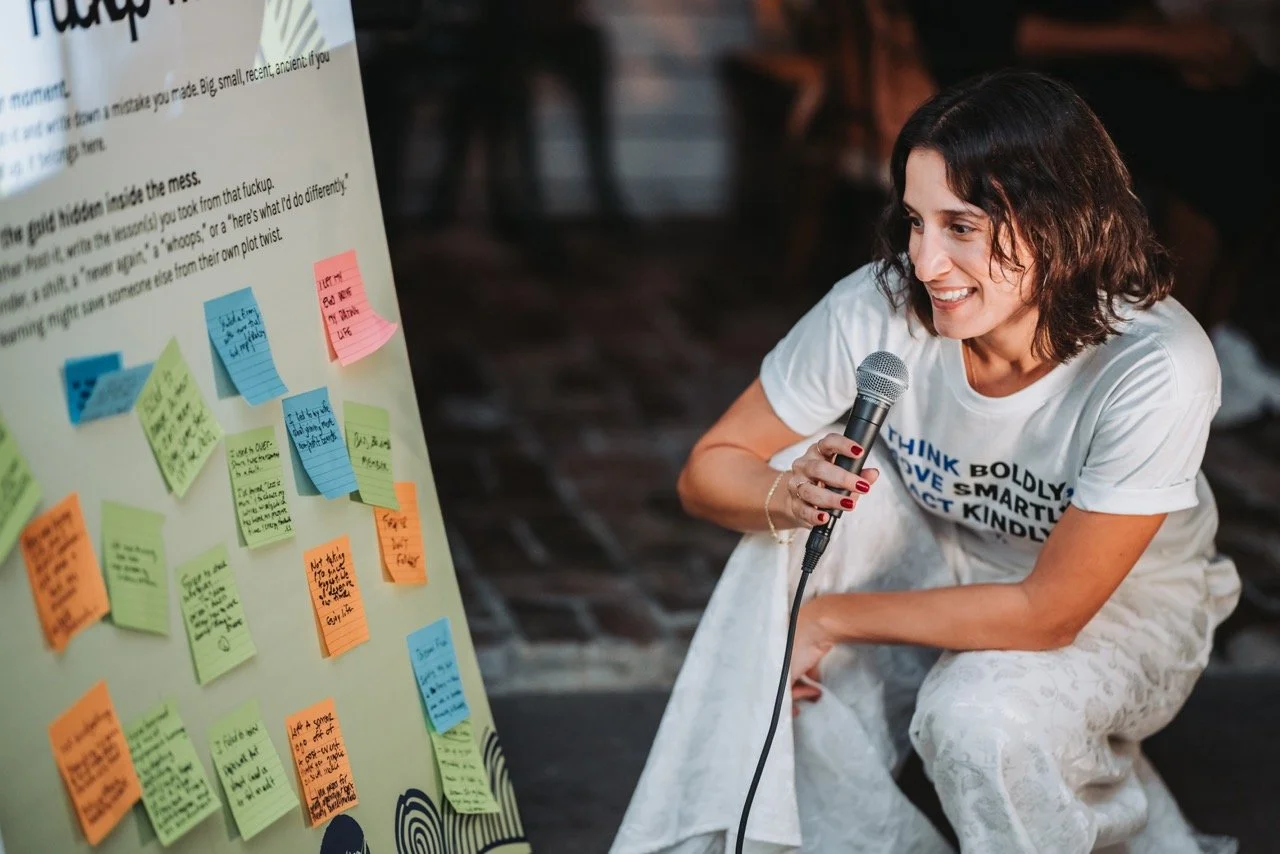 Alia Mahmoud holding a microphone, squatting next to a large whiteboard filled with colorful sticky notes.