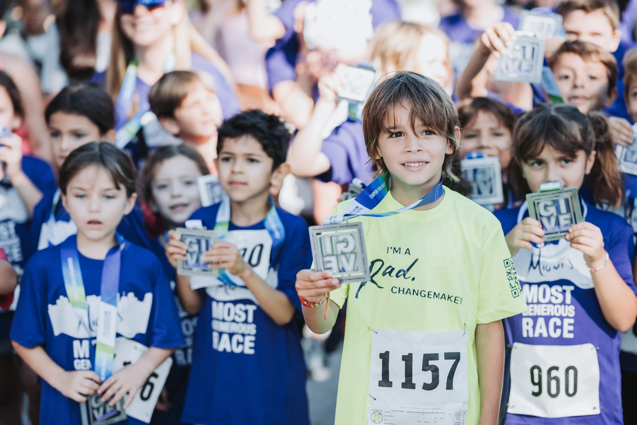 Children at a race event holding medals, some wearing blue shirts with race details, and a boy in a bright yellow shirt with the message 'I'm a Dad, Changemaker' and race number 1157 in the foreground.