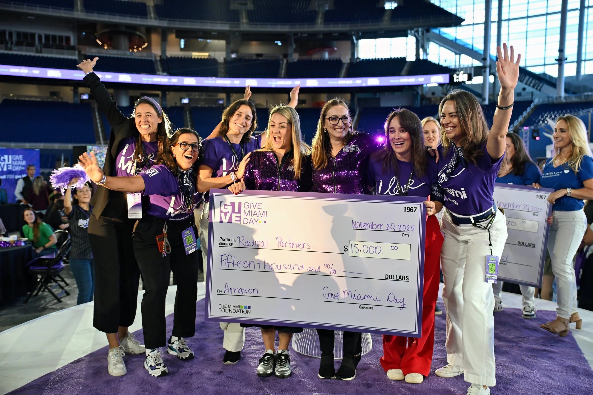 Group of women smiling and celebrating on stage at an event, holding a large check for $15,000 made out to Amazon, dated November 20, 2025, during Give Miami Day.