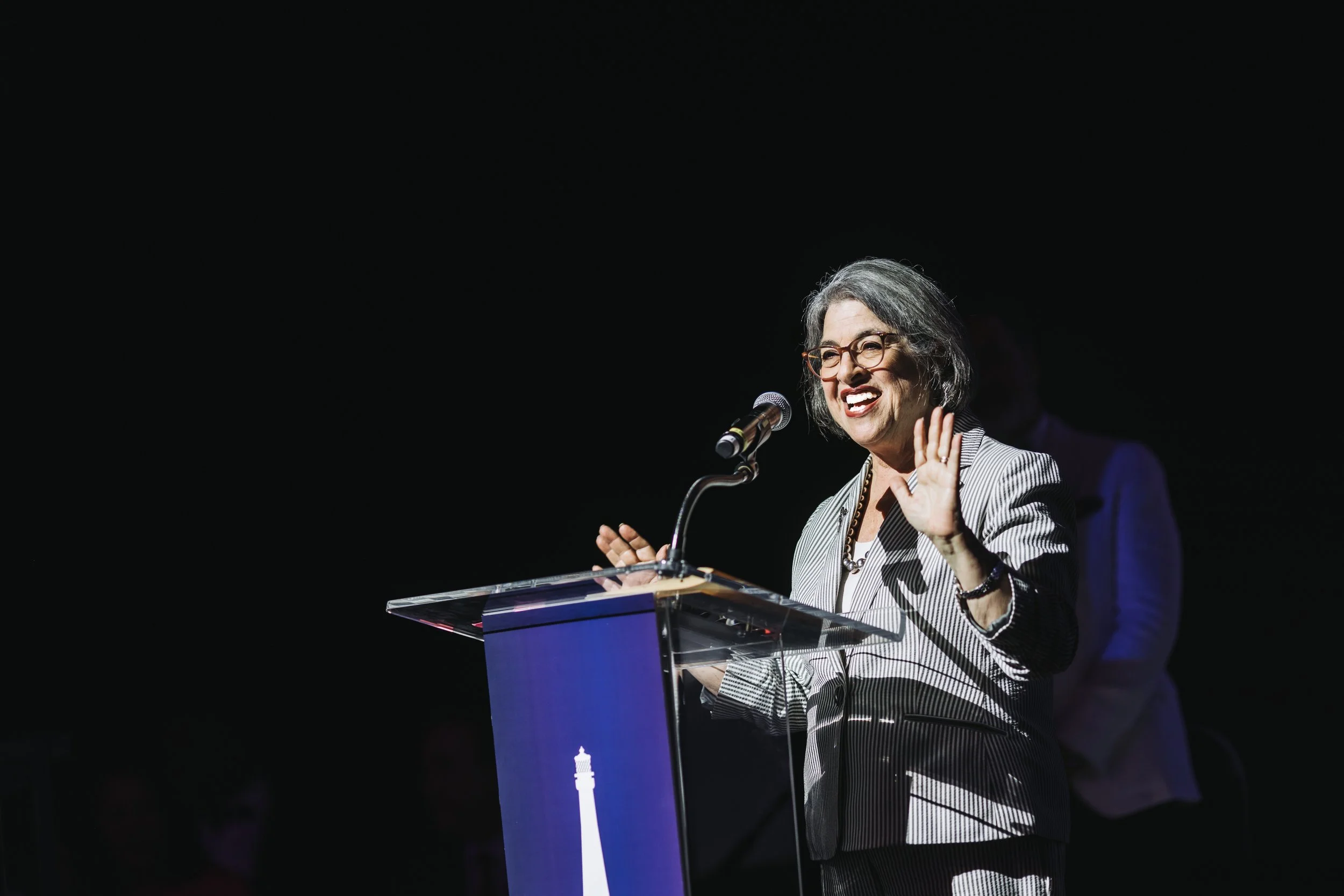 A woman with gray hair and glasses speaking at a podium during a public event, gesturing with her right hand and smiling.