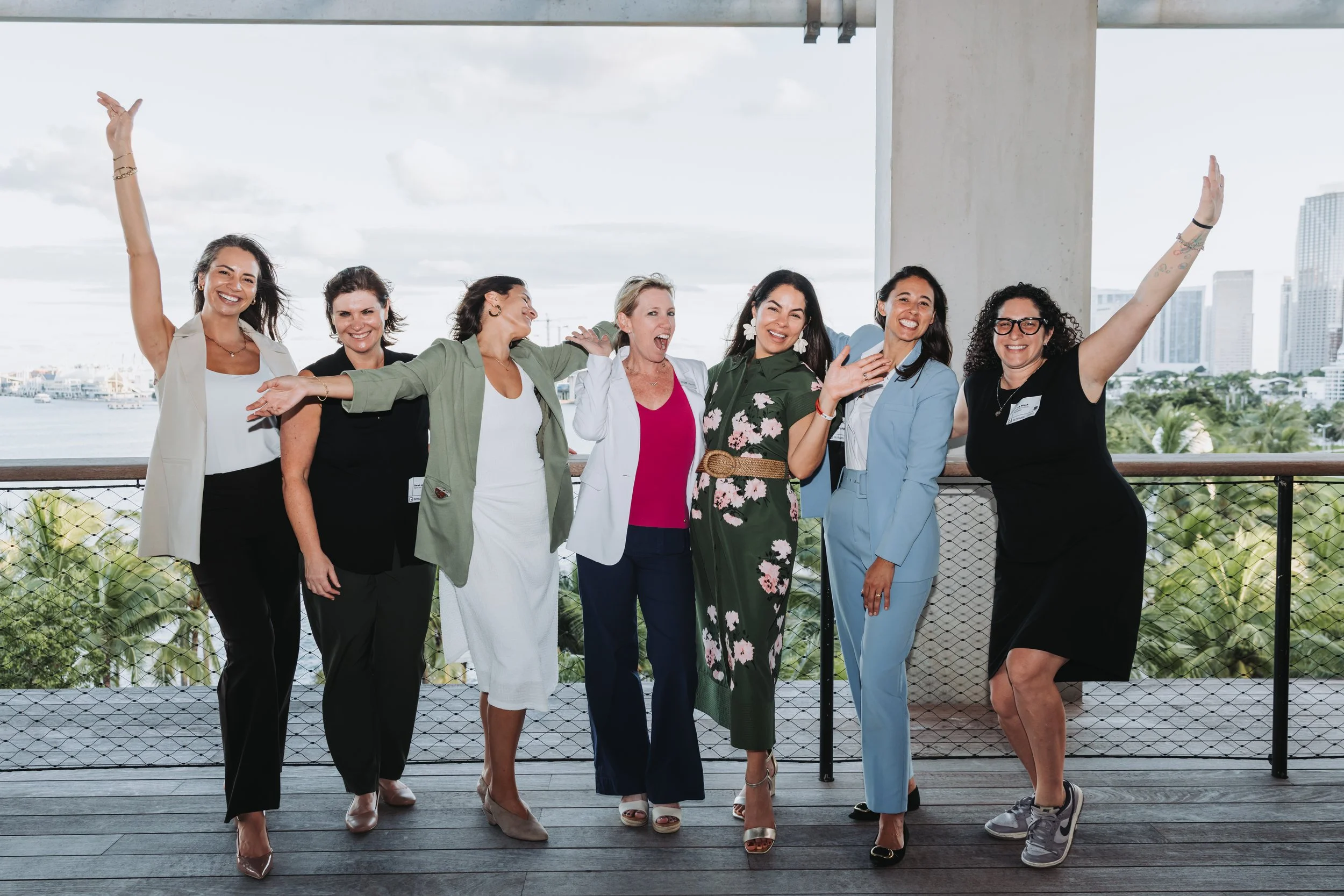 Seven women standing together outdoors, smiling and celebrating, with a city skyline and water in the background.