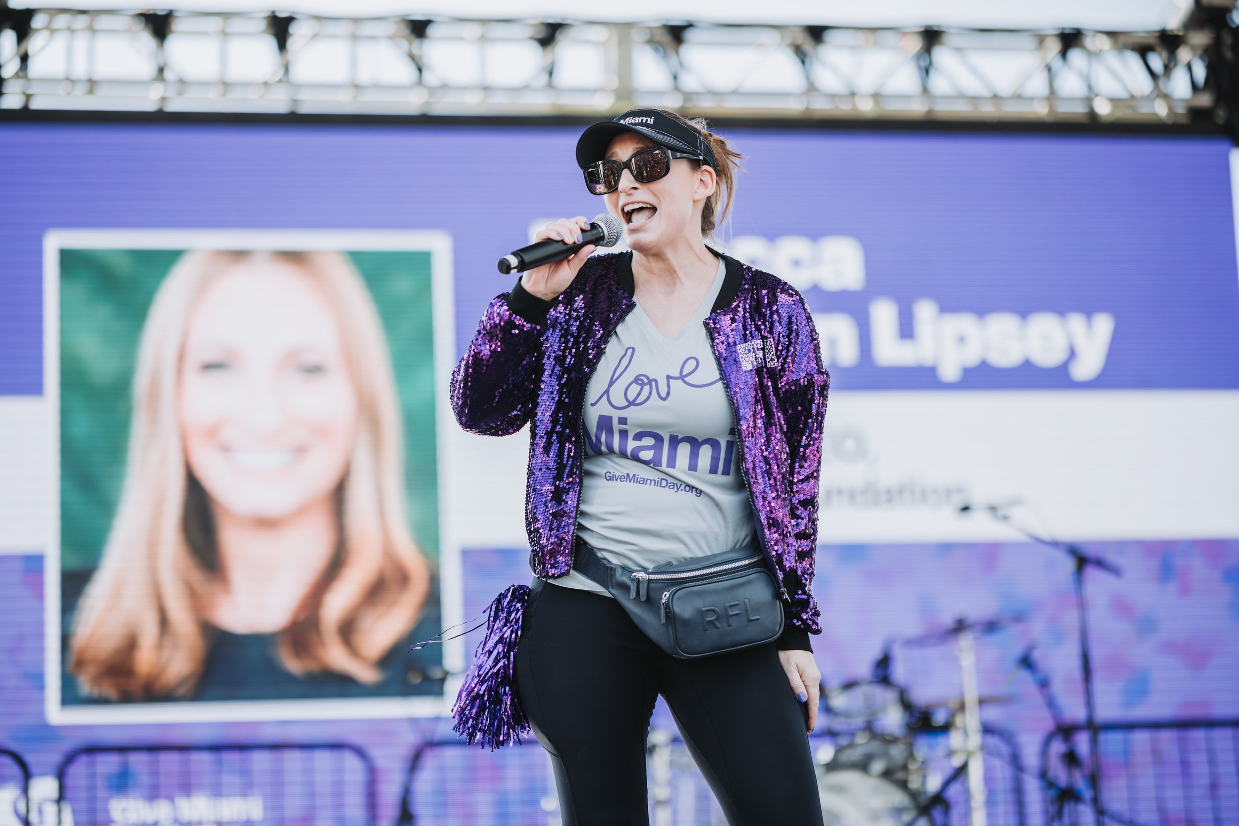 Woman speaking into a microphone on stage, wearing sunglasses, a black cap, a purple sequin jacket, and a gray t-shirt with 'love Miami' printed on it, at a public event or rally with a large digital screen in the background displaying her photo and 