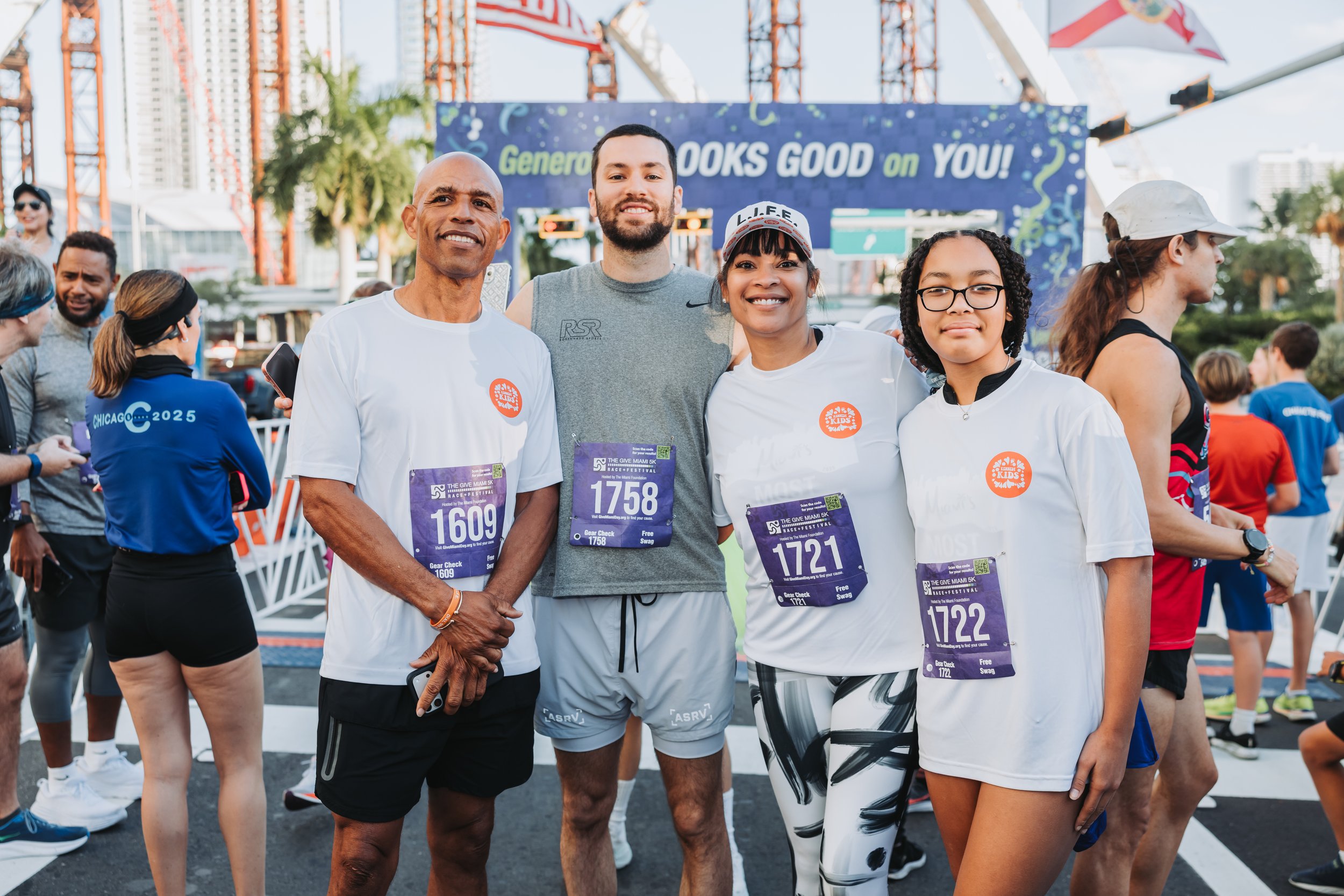 Four diverse runners with race bibs standing together at the starting line of a race with a carnival ride and people in the background.