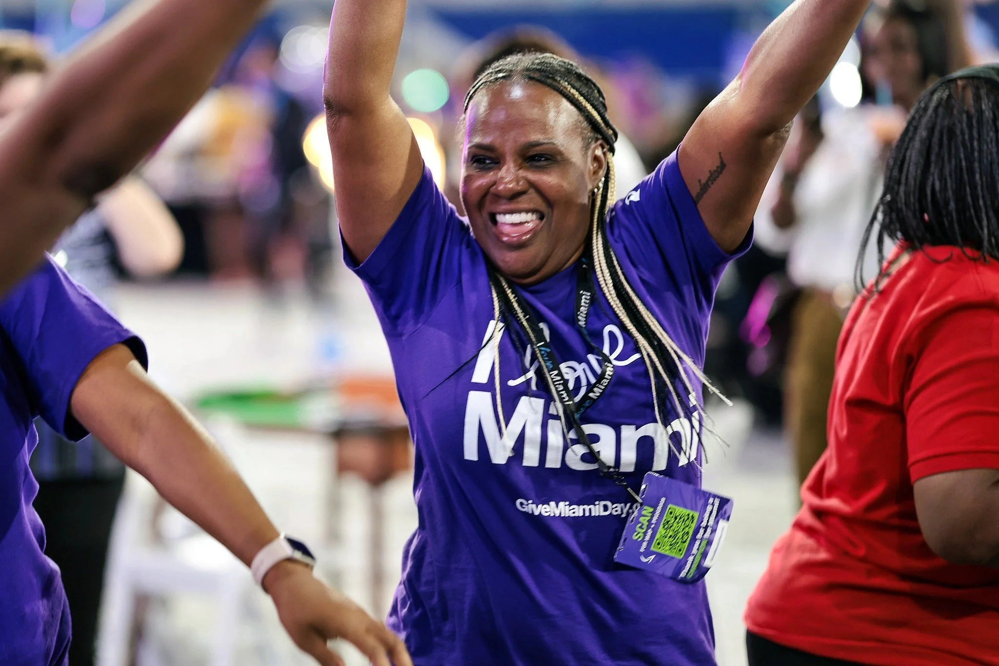 Smiling woman with braided hair in a blue shirt with 'Miami' printed on it, raising her arms in celebration at an event with many people in the background.
