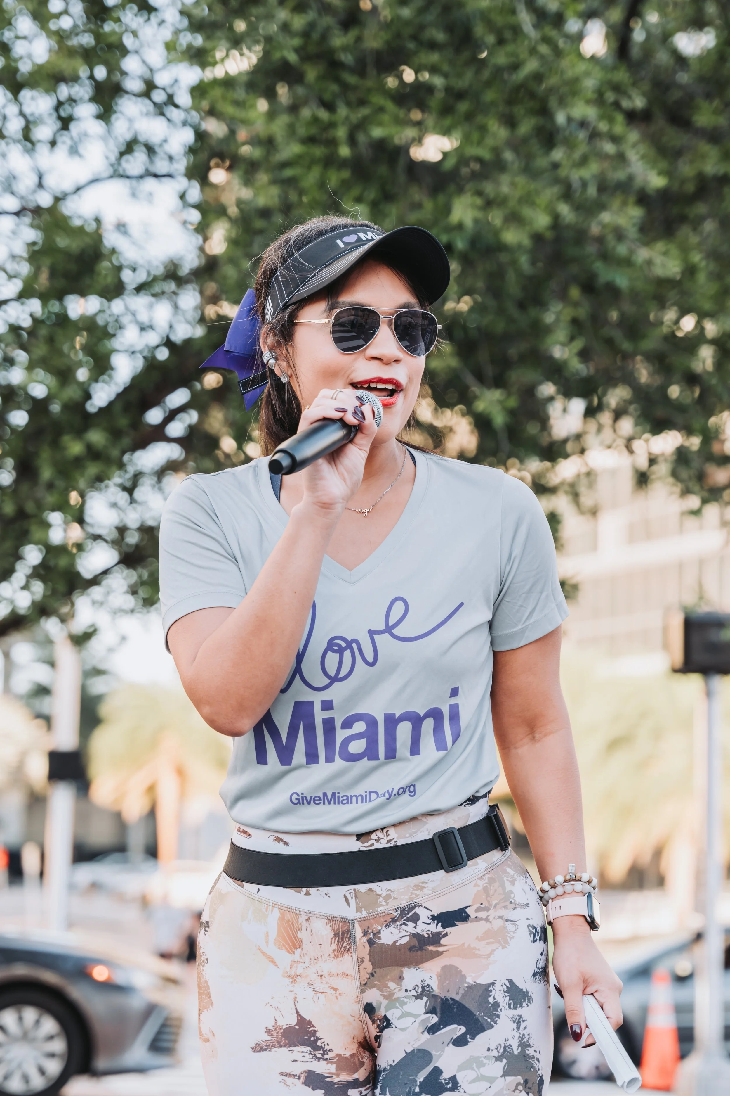 A woman wearing sunglasses, a black visor, and a gray t-shirt that says 'Love Miami' holding a microphone, standing outdoors with green trees in the background.