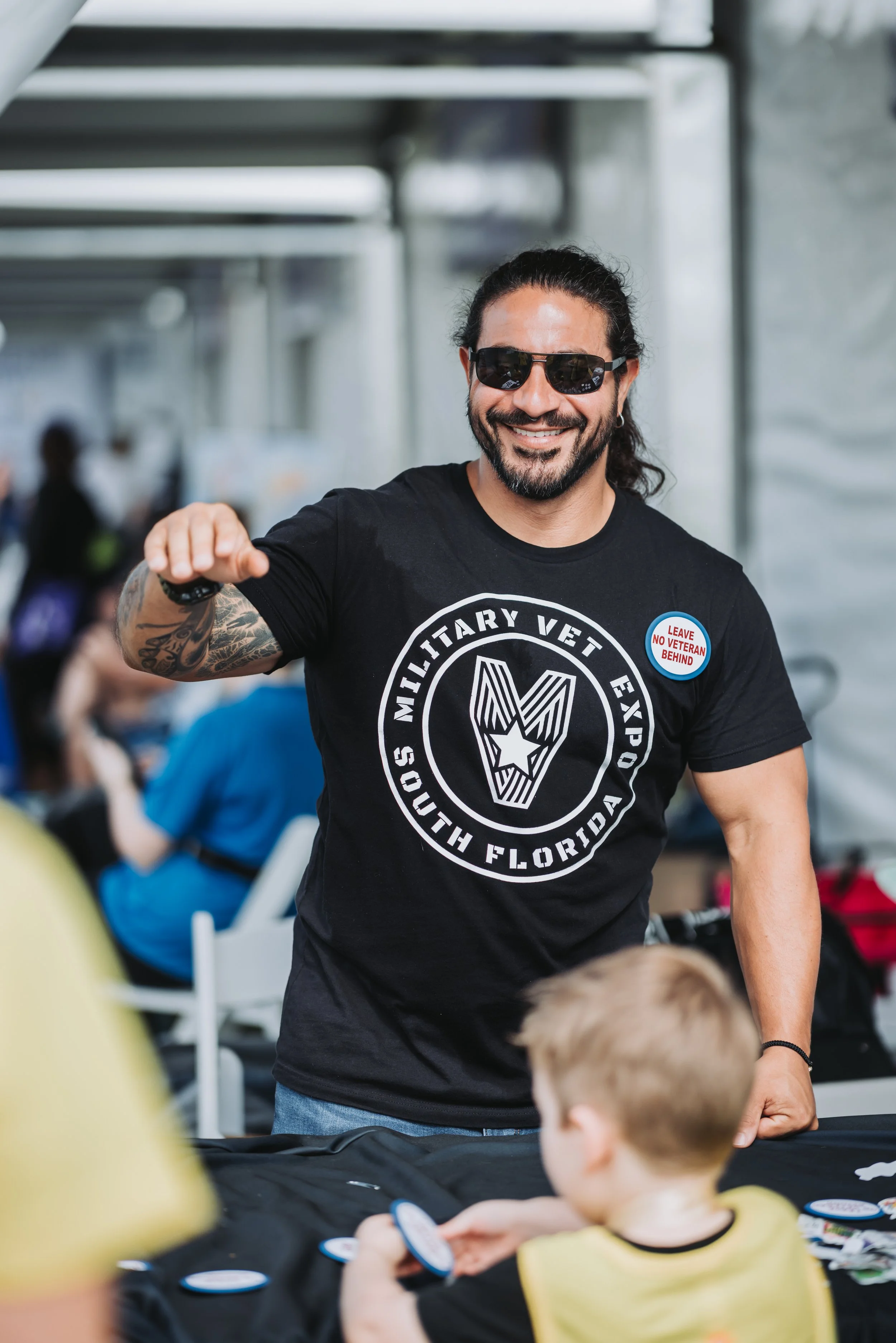 A smiling man wearing sunglasses, a black t-shirt with a military vet logo, and a badge that says 'Leave No Veteran Behind.' He is engaging with children at an event.