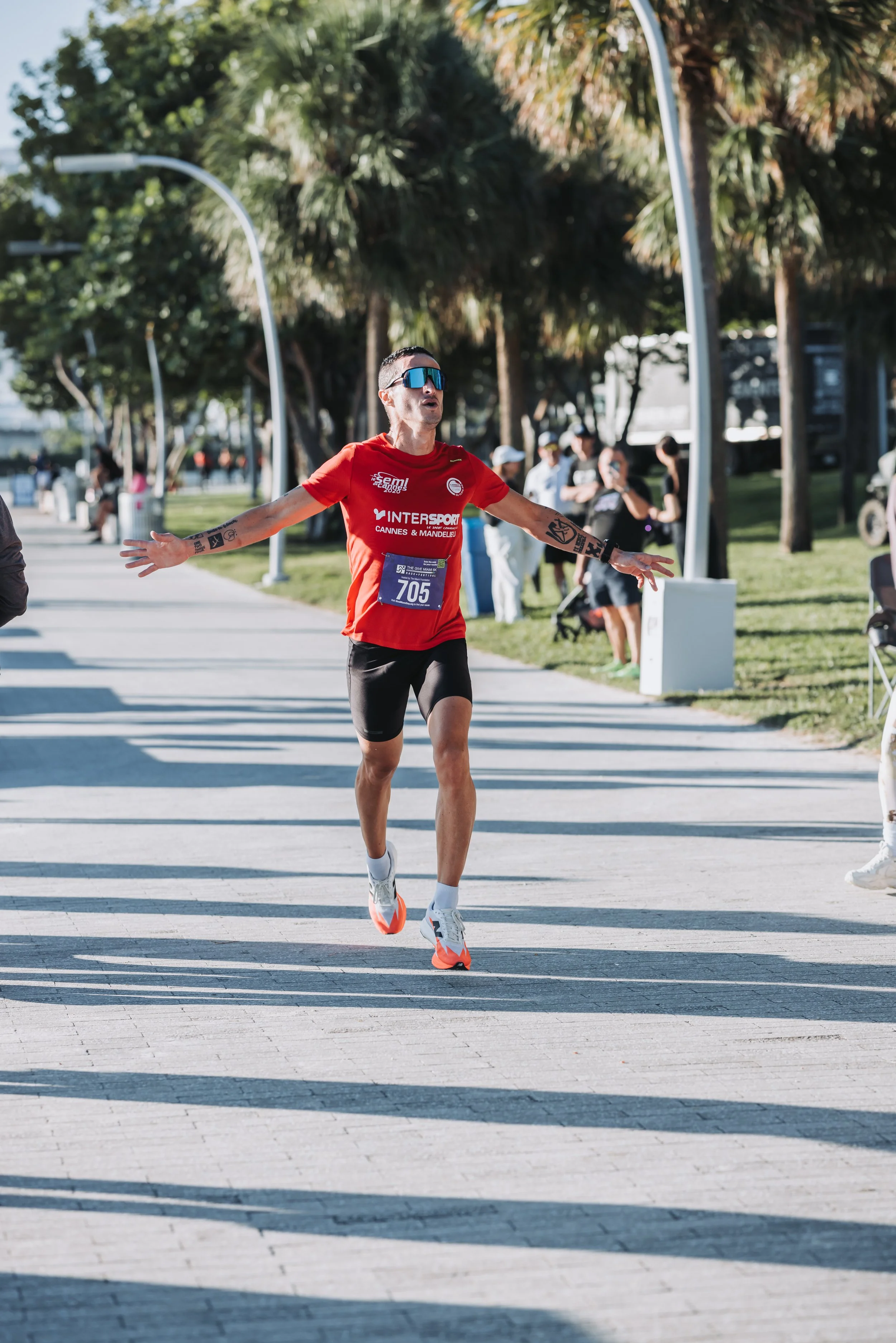 A male marathon runner in a red shirt and black shorts crossing the finish line with arms outstretched in a park-like setting with palm trees and spectators in the background.