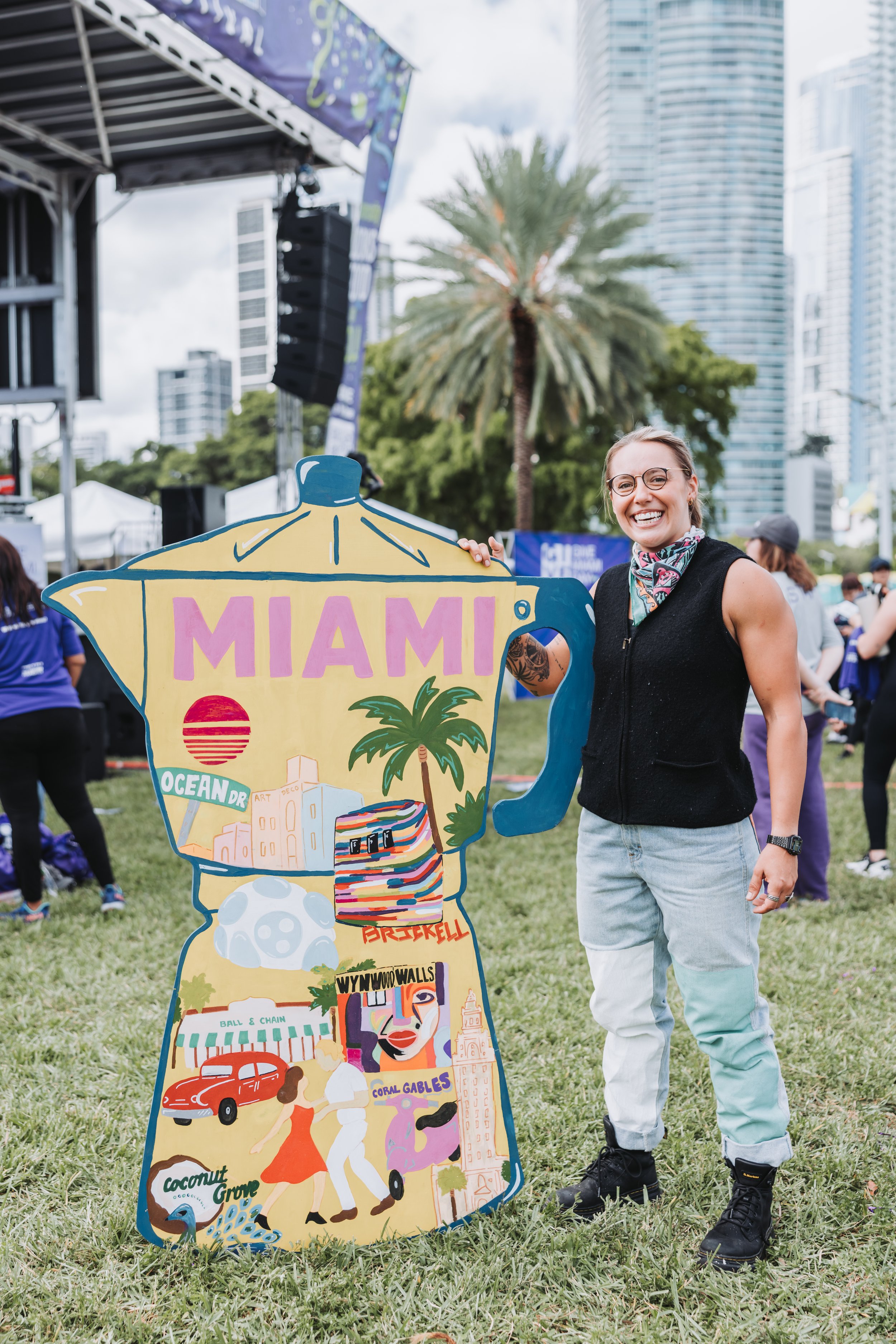 A smiling woman with glasses and a sleeveless vest stands outdoors on grass, holding a large, colorful cutout or sign shaped like a coffee pot with the word 'MIAMI' and illustrations of palm trees, buildings, and the Miami cityscape, during a daytime