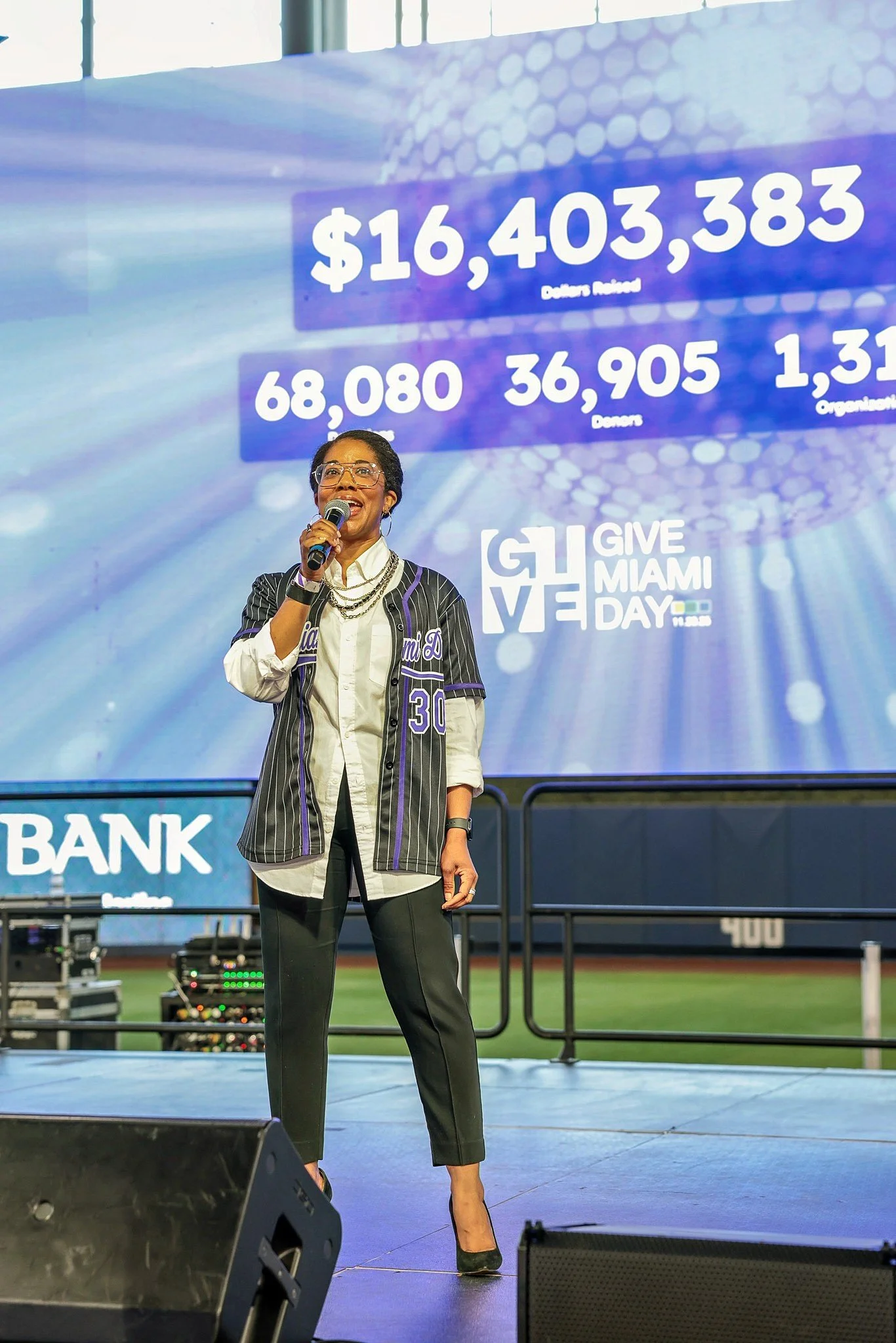 Woman in a baseball jersey speaking into a microphone on a stage with a large screen behind her displaying donation numbers and the event logo 'Give Miami Day'.