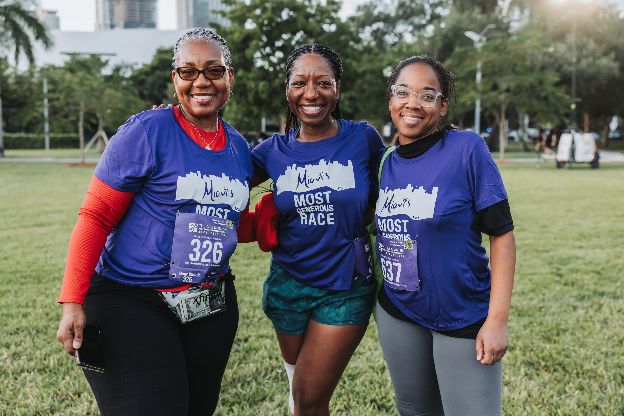 Three women wearing blue race shirts and bibs, smiling and standing outdoors in a park during daytime.