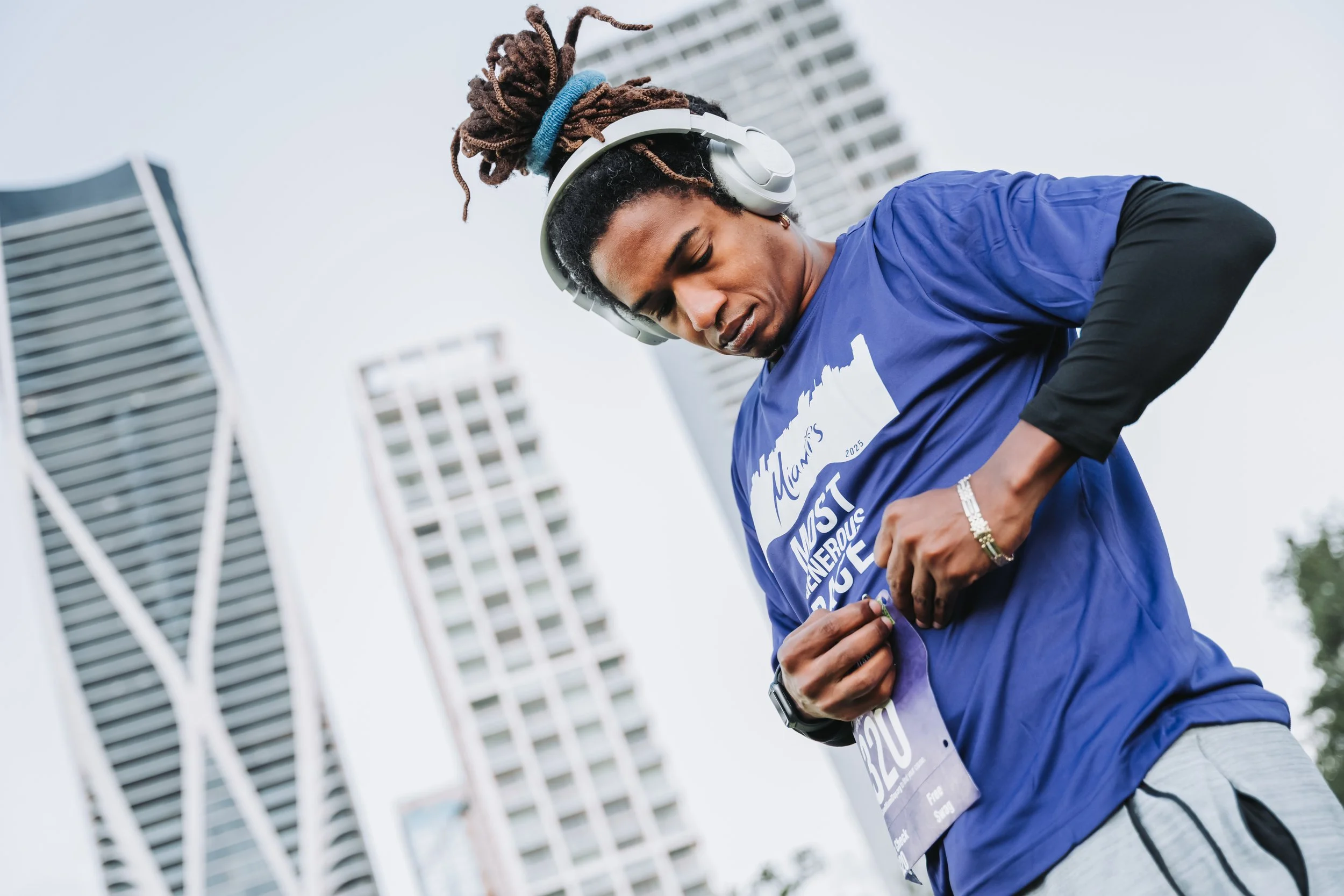 A man with dreadlocks and headphones finished a race, wearing a blue shirt with event-related text, standing in an urban area with tall buildings.