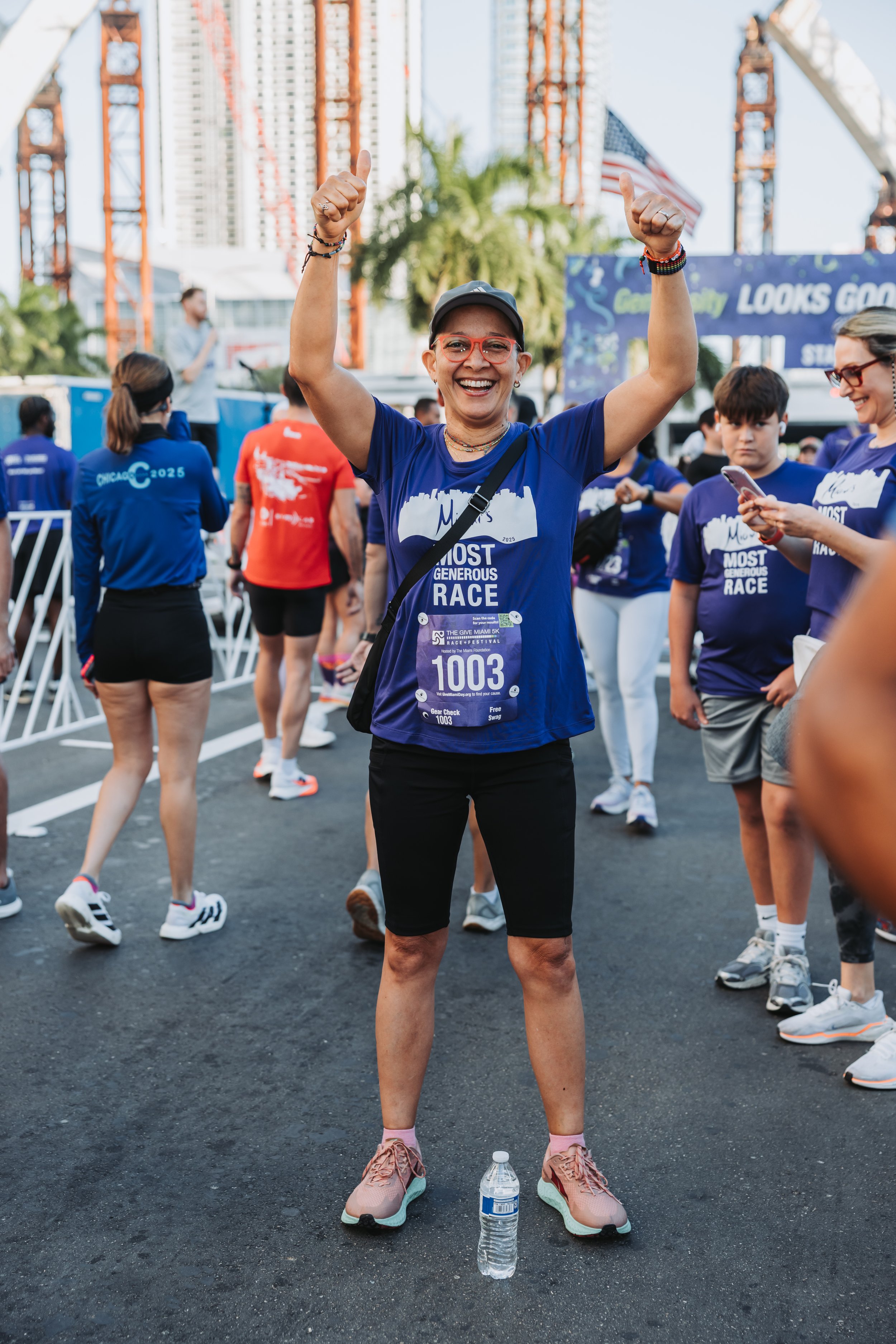 A woman smiling and raising her arms in victory at a running event, surrounded by other participants, with city buildings and cranes in the background.